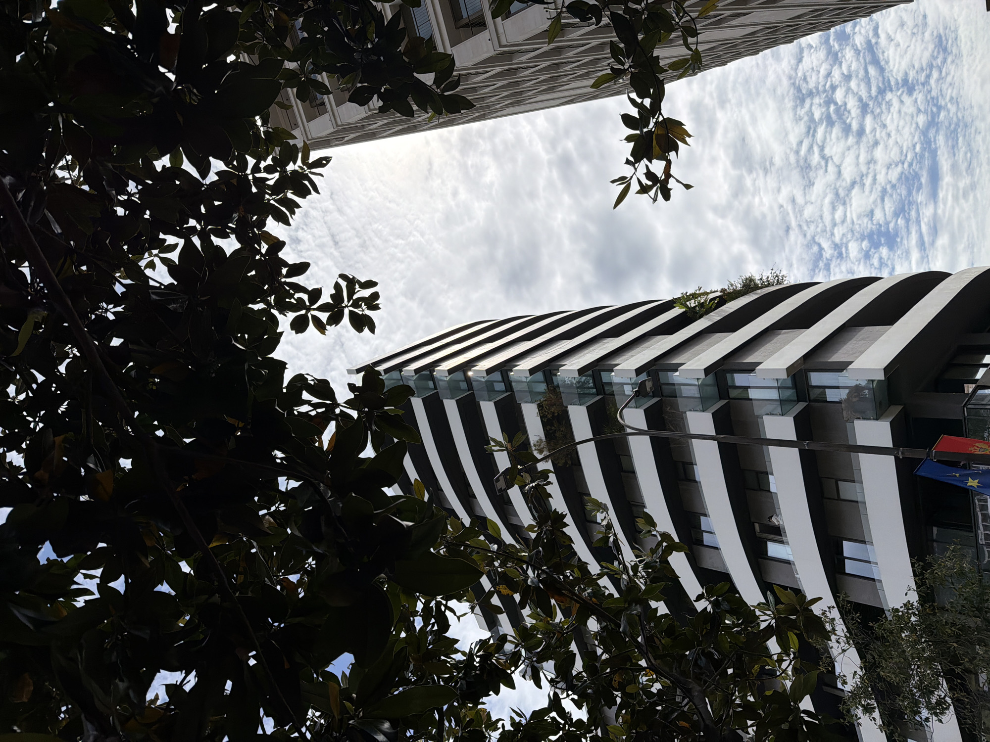 The low-angle shot captures two buildings and the sky through foliage. One building features a modern design with alternating white and dark horizontal stripes. The other building is a taller structure with a textured facade. The sky is filled with scattered clouds. Leaves frame the scene in the foreground. A flagpole with flags is visible on the striped building. This is a view of buildings in Budva, Montenegro.