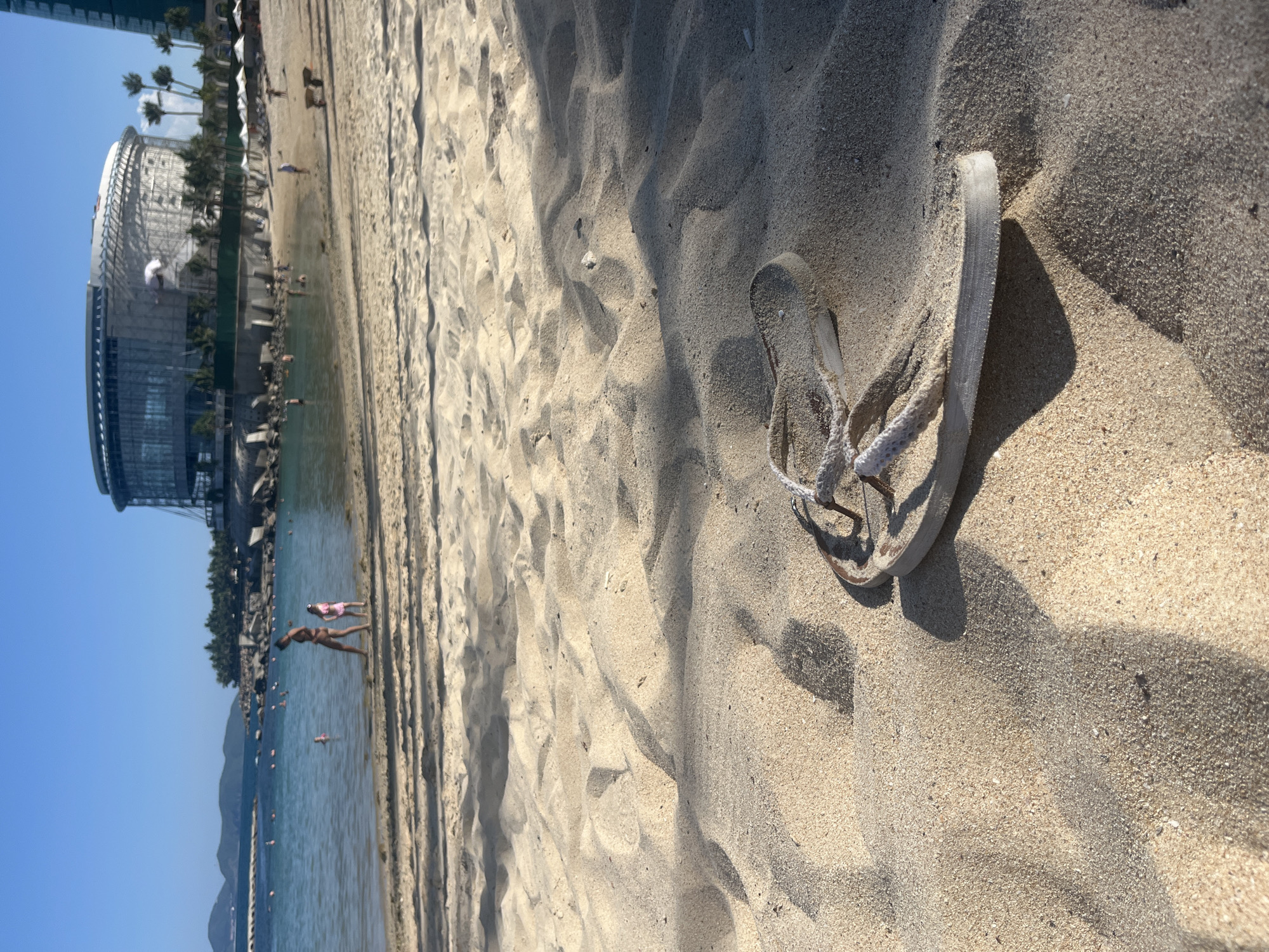 A close-up, low-angle shot focuses on a single white flip-flop resting in rippled sand on a beach. In the middle ground, a woman in a bikini and a young child in a pink swimsuit walk near the water's edge. The sea is calm with several people swimming in the distance. A breakwater made of large rocks stretches into the water, and beyond it, a large, futuristic-looking circular building with glass panels and a domed roof is visible. Palm trees and other vegetation are scattered around the building, and a few white beach umbrellas are set up on the sand to the right. The sky is clear and blue.