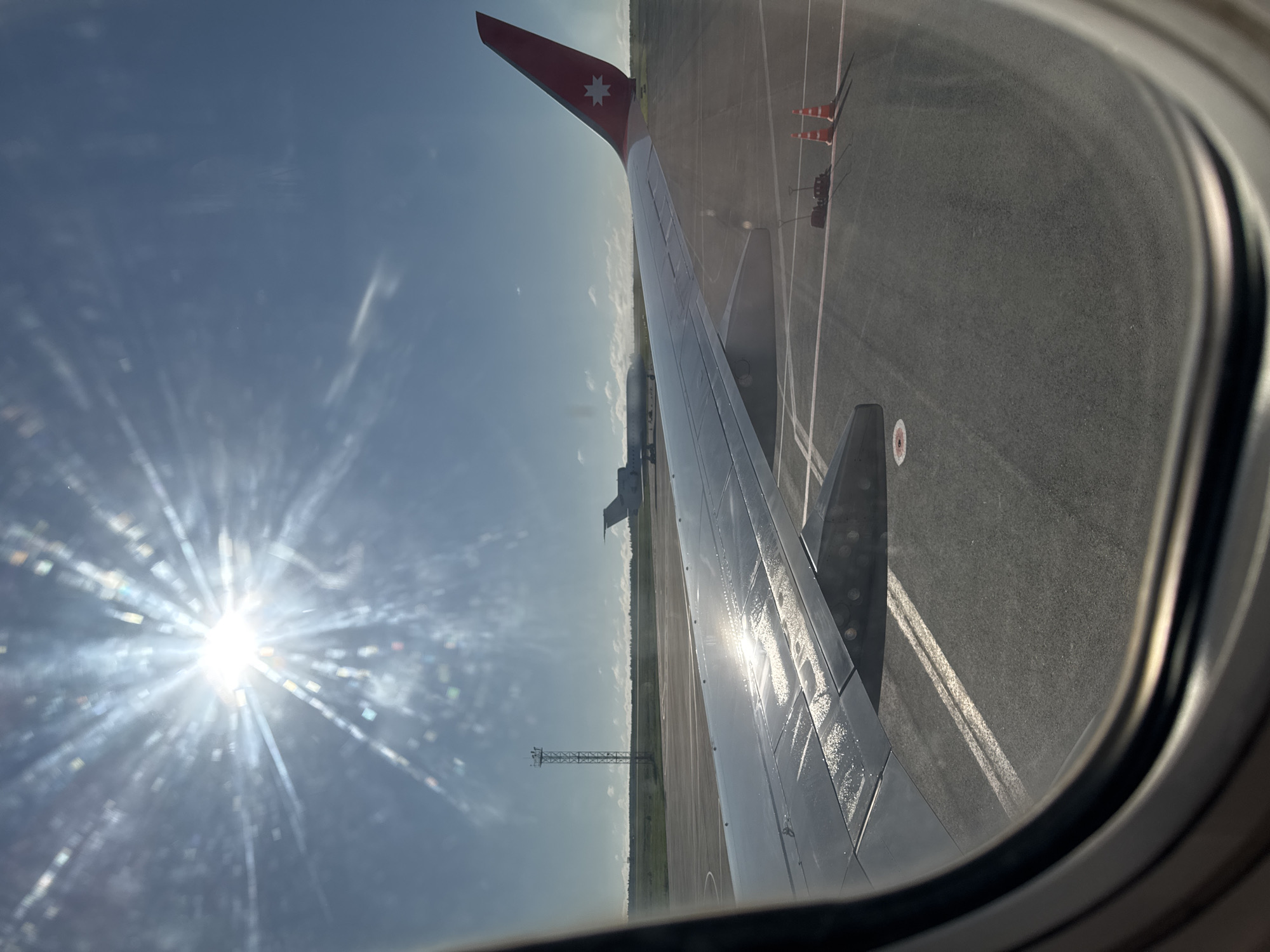 This is a photograph taken from an airplane window, looking out at an airport tarmac. The sun is bright and appears to be setting or rising, casting lens flare across the image. In the foreground, a portion of the airplane's wing is visible, with its red and white tail fin featuring a white star logo. The wing is wet, reflecting the sunlight. In the background, another airplane is parked on the tarmac, and a tall antenna structure stands to the left. The tarmac is paved and marked with white lines. Two orange traffic cones and some equipment are also visible on the ground. The sky is blue with some scattered clouds. The image is framed by the curved edge of the airplane window. There are no people visible in the image. The location is identified as Staroe Mart'yanovo, Russia, implying this is an airport in or near that settlement.