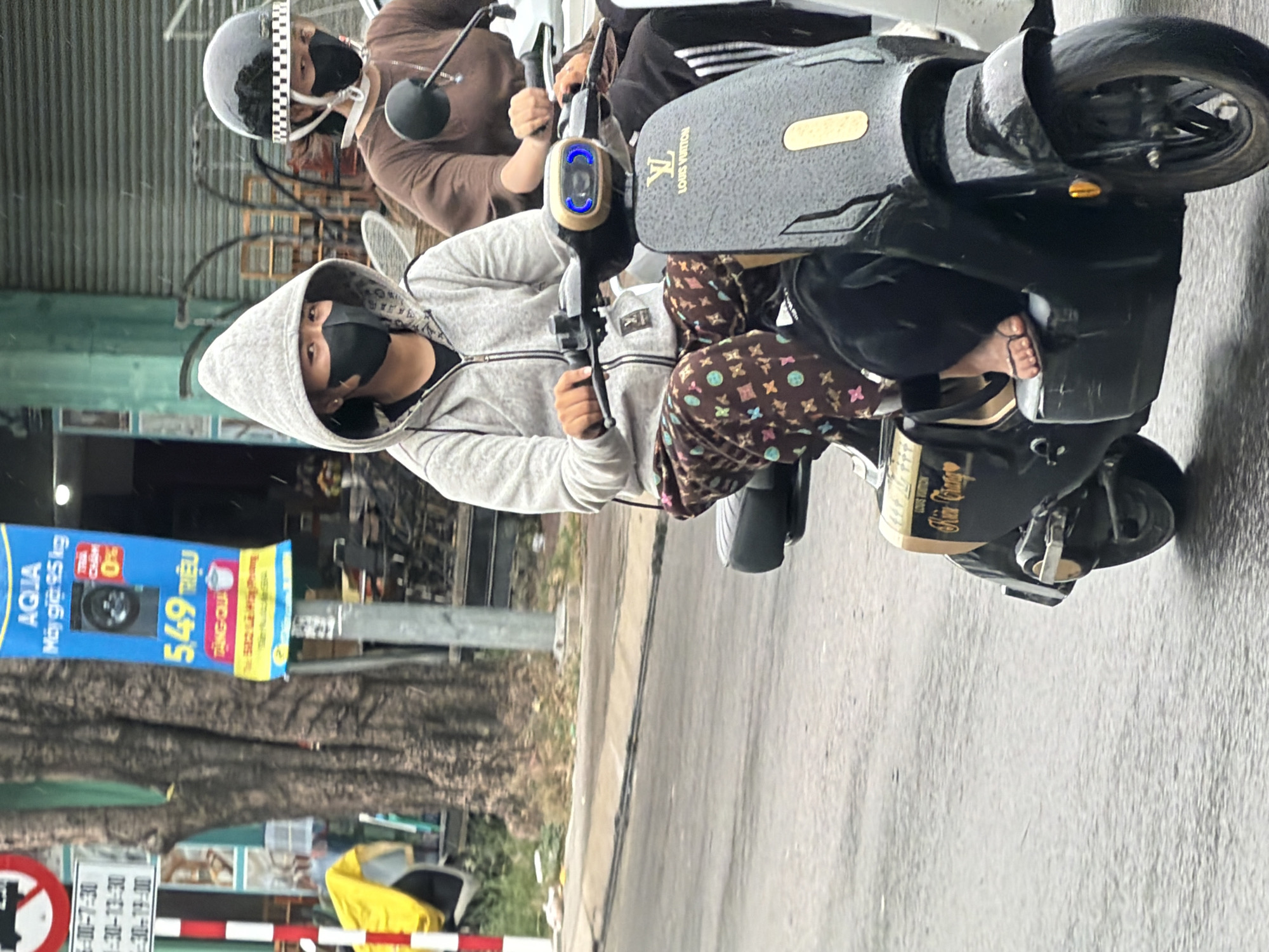 In Nha Trang, Vietnam, two individuals are riding on separate electric scooters on a wet, paved road. The person in the foreground, wearing a grey hooded jacket, black face mask, and patterned pants, is operating a black scooter adorned with the Louis Vuitton logo. The scooter has blue accent lights on the handlebars. The second person, riding slightly behind and to the right, is wearing a brown shirt and a black helmet with a checkered band. The environment appears to be an urban street with trees, a pole with signage, and buildings in the background. The weather seems overcast, and the road surface is reflective, indicating recent rain.