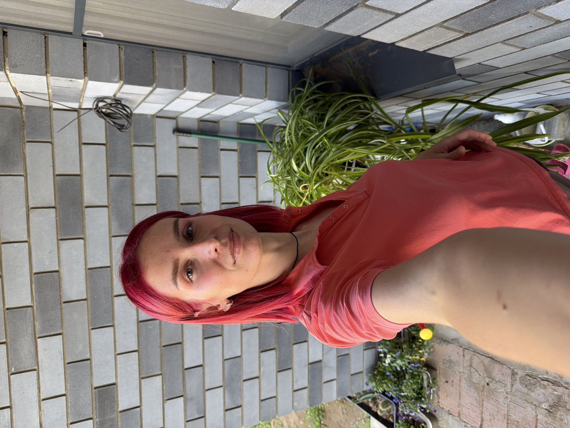 A female individual, approximately 20-30 years old, with bright magenta-red, shoulder-length hair, is depicted taking a selfie outdoors in Chaikovsky, Russia. She has fair skin, light-colored eyes, and wears minimal visible makeup, with small hoop earrings and a thin black choker necklace. Her attire consists of a coral-pink short-sleeved polo shirt with snap buttons. She maintains a neutral expression with a slight smile, gazing directly at the camera, which is held at arm's length, likely by her right hand, revealing a small mole and a faint scratch on her arm.
The background features a multi-toned grey concrete block wall, laid in a running bond pattern, indicating the exterior of a building. To the immediate right, a dark grey window or door frame is partially visible. A coiled bundle of dark electrical wire is affixed to the upper right section of the wall. At ground level, behind the woman, a large spider plant with variegated green and yellow leaves grows in a white pot. The green handle of a garden tool leans against the wall behind this plant. To the left, a rectangular planter contains small cacti and flowering plants with purple and yellow blossoms. The ground visible to the lower left consists of light-colored pavers or bricks, transitioning to an area of bare earth or grass. A small yellow spherical object is partially visible on the ground behind the woman's arm. The scene is illuminated by natural daylight.