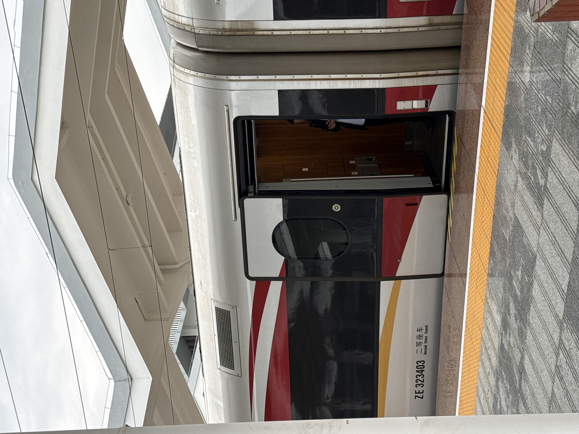 The image shows a train car at a station in Yongding District, China. The train car is painted white, black, and red. The number "ZE 323403" and the words "Second Class Coach" are printed on the side of the train car. The train door is open. A person is inside the car. The station platform is made of gray stone with a yellow tactile warning strip. A building with a geometric architectural style stands next to the train station.