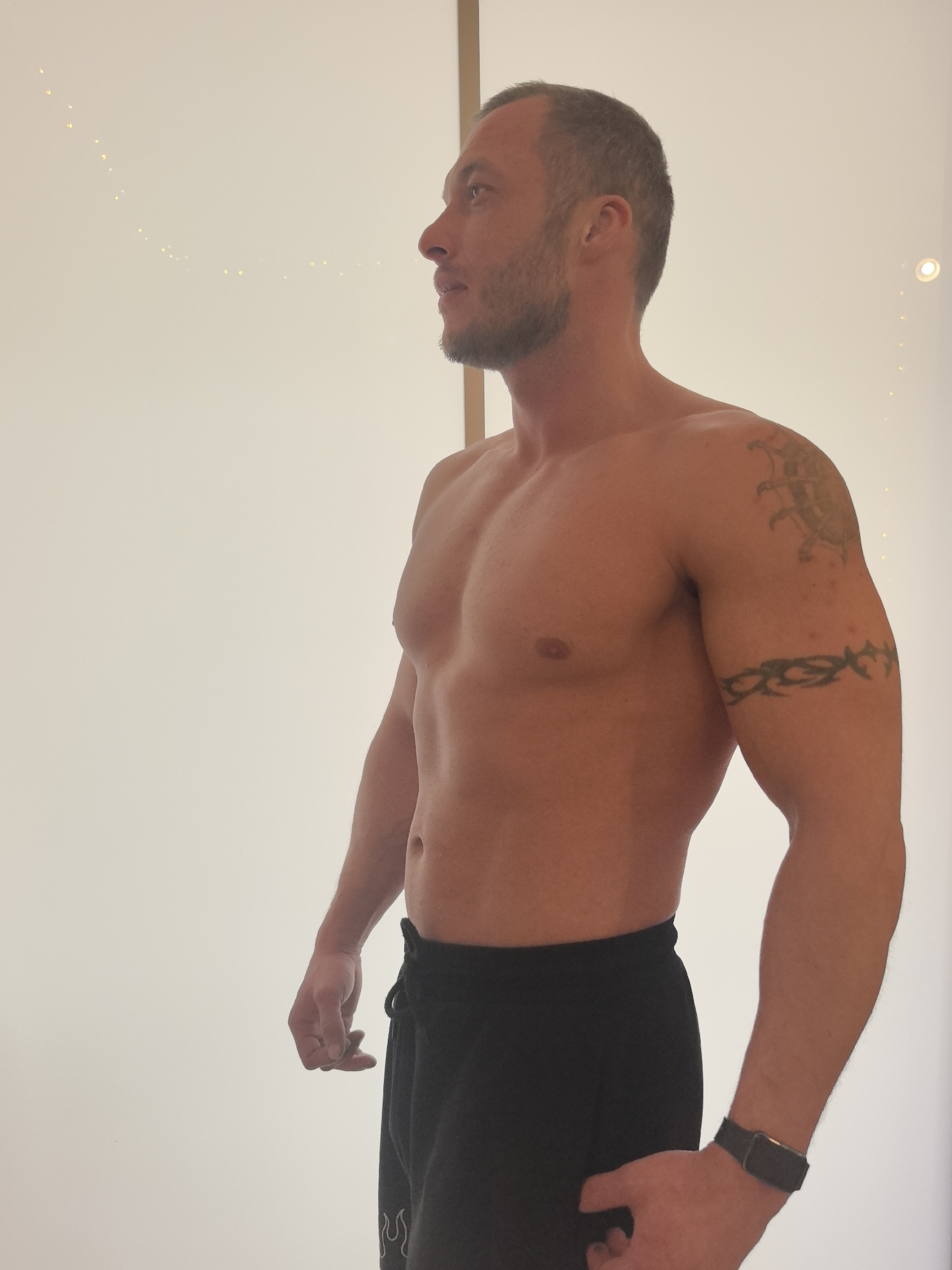 A shirtless man is standing indoors, facing left. He has a muscular physique, with a chest and arm tattoo. He is wearing black shorts and a fitness tracker on his left wrist. The background is a plain white wall with a string of lights and a single spotlight. The man is not in Nizhny Novgorod, Russia.