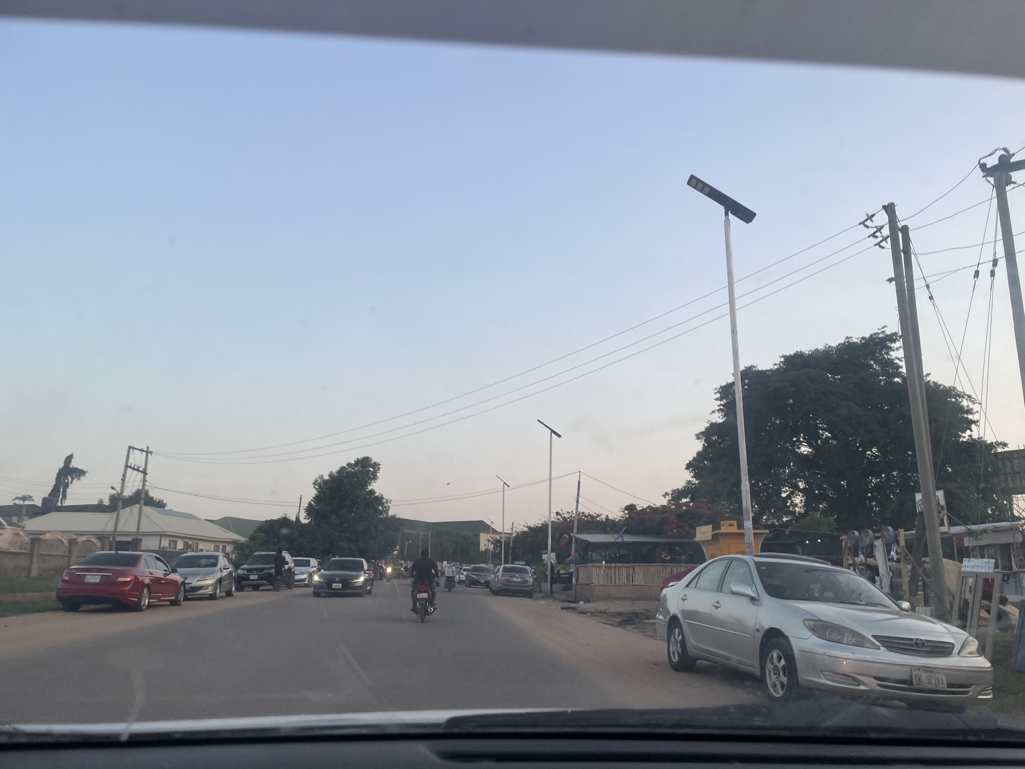 The image depicts a street scene in Abuja, Nigeria, during daylight hours. A silver Toyota Camry is parked on the right side of the road. The road is asphalt and has traffic flowing in both directions, including cars and a motorcycle. Street lights line the road. Power poles and wires are visible. Buildings and trees are in the background.