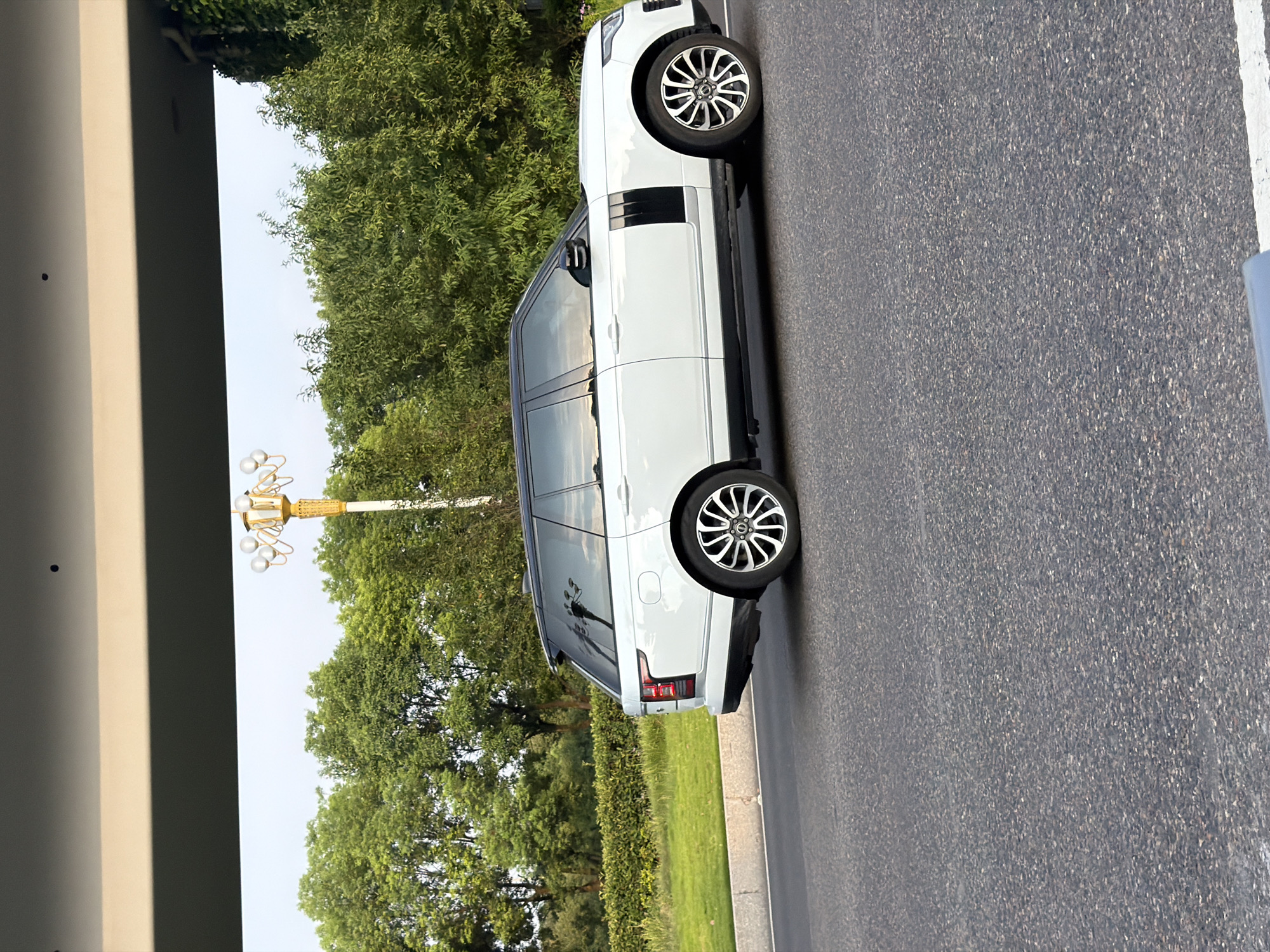 A white Range Rover is driving on a paved road in the city of Jiangjin, China. The car is moving from left to right in the frame, and its side is visible. The car is in good condition and appears to be a recent model. The road is asphalt and appears to be in good condition. There are trees and bushes on the side of the road, and a street light is visible in the background. The sky is overcast, and the overall lighting suggests it is either early morning or late afternoon. The scene is a typical urban or suburban setting.