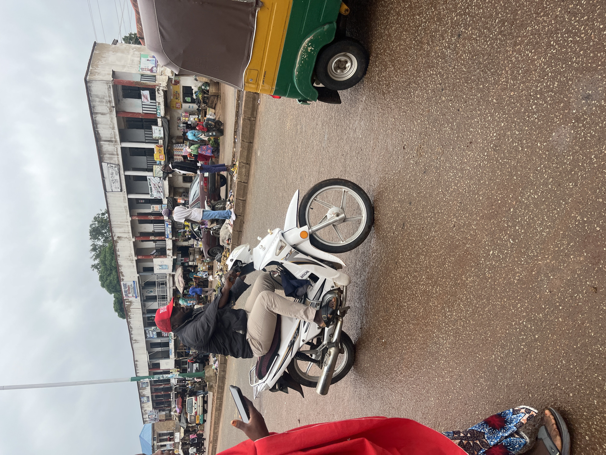 In Bukuru, Nigeria, a man wearing a red baseball cap and a black jacket rides a white motorcycle on a wet asphalt road. He is wearing light-colored pants and sandals. The motorcycle has a silver exhaust pipe and black detailing. To the right of the motorcycle, a yellow and green auto-rickshaw is parked. In the background, a row of buildings with shops line the street. People are visible walking and standing in front of the shops. The sky is overcast. To the left of the motorcycle, a person in a red garment and patterned fabric is partially visible, holding a smartphone.