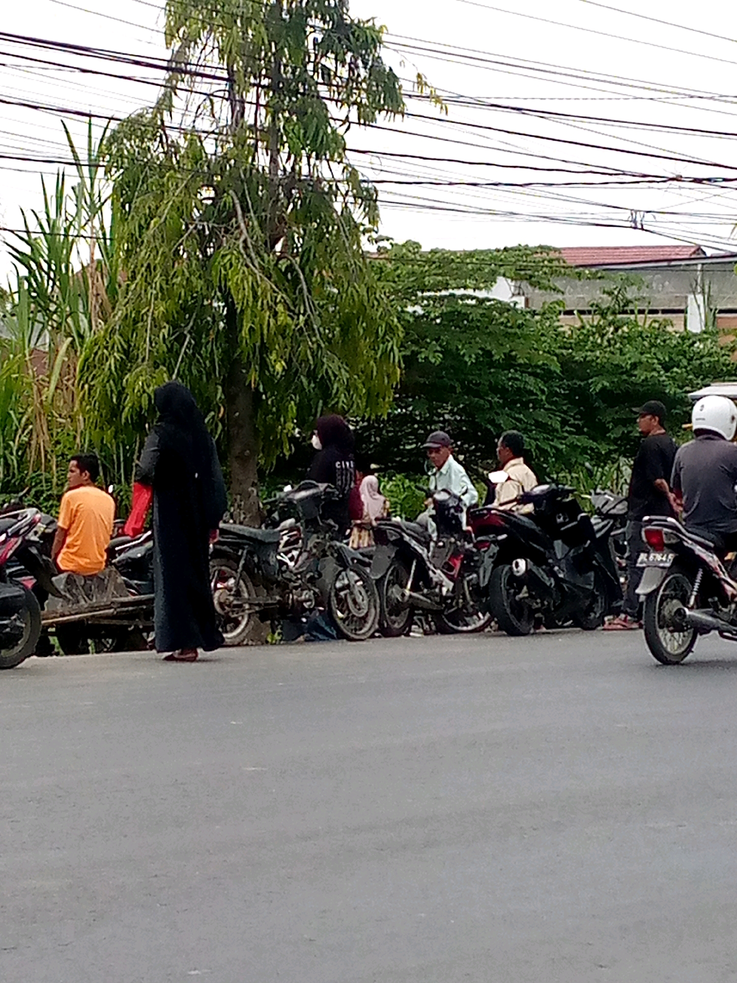 The image depicts a street scene in Keudebayu, Indonesia, likely during the daytime. The foreground features a grey asphalt road. To the left and center, a line of parked motorcycles is visible, positioned alongside a raised wooden barrier or sidewalk. Beyond the motorcycles, a dense arrangement of trees and foliage forms a natural backdrop. In the background, buildings with red-tiled roofs and other structures are partially obscured by greenery. Several individuals are present, some seated on or standing near the motorcycles, while others are walking. One woman in a black hijab and long black dress is walking on the road in front of the motorcycles. Another woman in a black headscarf and dark clothing stands near the motorcycles. A man in an orange t-shirt is seated on one of the motorcycles. Several other men are also visible, some wearing hats and casual attire, standing and conversing near the parked vehicles. The overall impression is a typical roadside gathering in a tropical urban or semi-urban environment.
