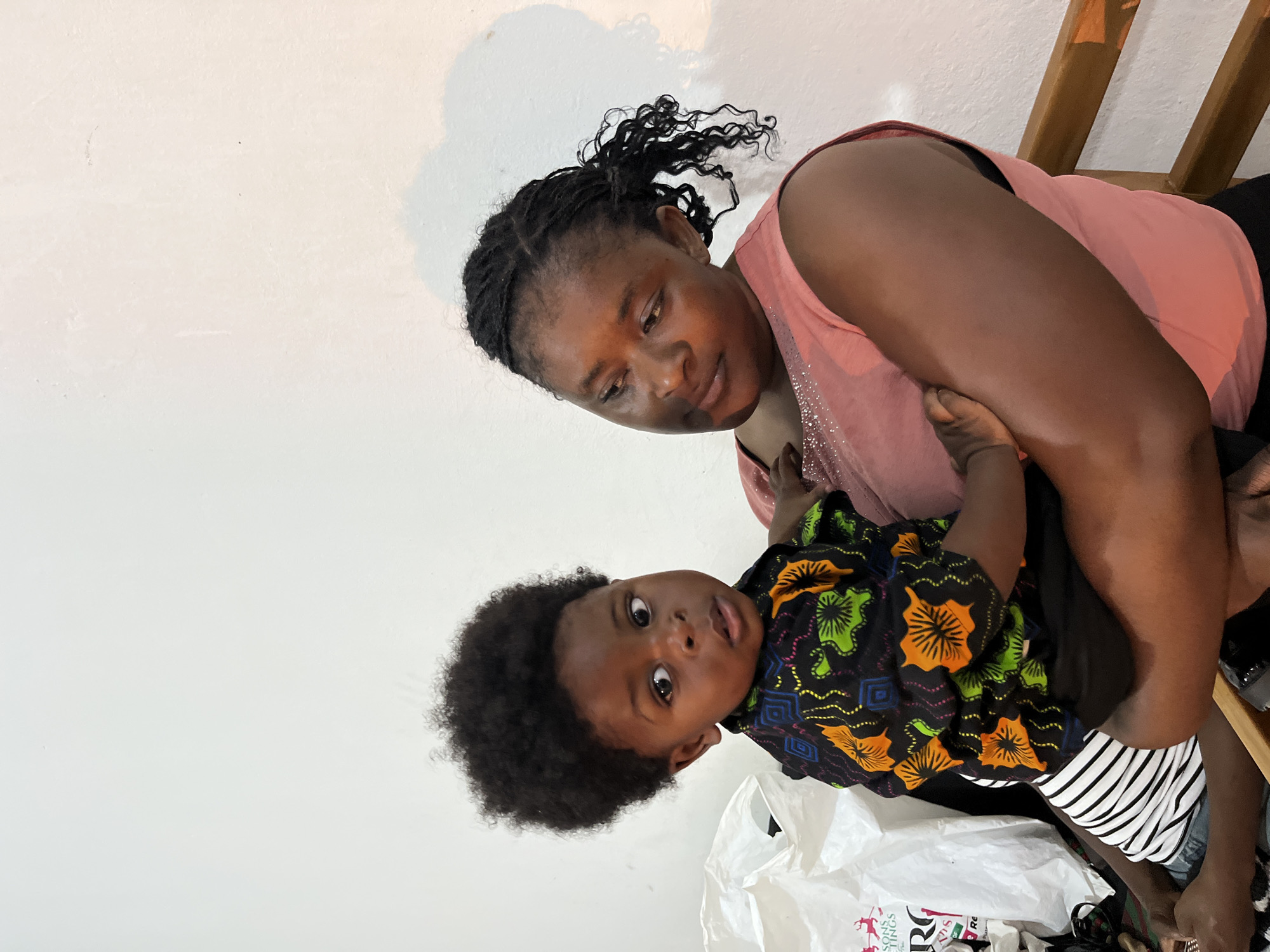 In a dimly lit indoor setting, a woman with dark braided hair, wearing a pink sleeveless top adorned with subtle sparkly embellishments, cradles a young child. The child, with voluminous dark curly hair, is dressed in a black short-sleeved shirt featuring a vibrant, patterned design of orange, green, and blue shapes. The child is also wearing a black and white striped garment underneath. The background consists of a plain white wall, with a subtle shadow cast on it, and a large white plastic bag is visible to the left of the subjects. A wooden railing is present in the lower right portion of the image. The scene appears to be in Gura Top, Nigeria.