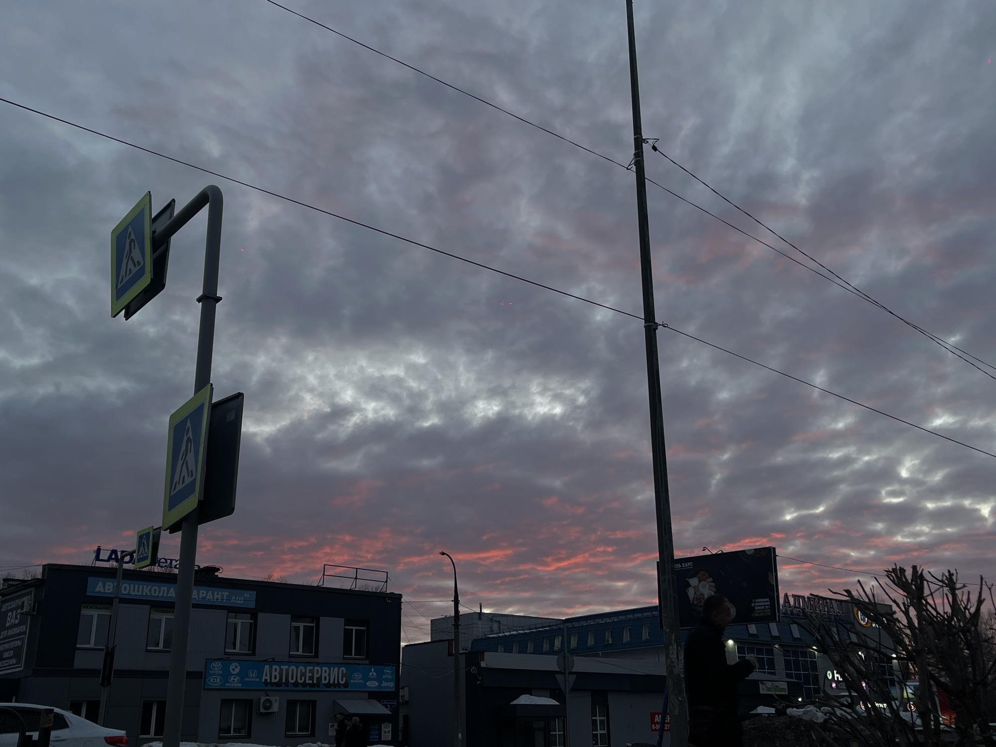 The image captures an urban street scene in Chaikovsky, Russia, at either dawn or dusk, under a sky characterized by a dramatic display of grey, orange, and red clouds. The upper sky is predominantly grey and overcast, transitioning to vibrant orange and red hues within the cloud formations closer to the horizon. Overhead power lines or utility wires stretch horizontally across the upper and middle portions of the frame.
In the left foreground, a grey pole supports two blue and white pedestrian crossing signs, one angled towards the viewer and the other perpendicular. A portion of a white vehicle is visible below these signs. Behind the pole stands a two-story building with a dark grey facade and multiple windows. This building features prominent signage, including "ЛАДА ДЕТАЛИ" (Lada Parts), "АВТОШКОЛА ГАРАНТ АШ" (Driving School Garant AS), and "АВТОСЕРВИС" (Auto Service) with car brand logos (Toyota, Kia, Hyundai, Opel, Skoda, Jeep, Subaru). Two indistinct figures are partially visible near the building's entrance on the ground level.
On the right side of the image, a tall, slender dark grey utility pole extends vertically through the frame, anchored with several horizontal wires. To the right of the image center, a silhouetted individual, dressed in a dark jacket, stands facing left with their right arm slightly raised. Immediately adjacent to this person is a dense cluster of bare, dark branches, possibly from a pruned shrub or tree. In the midground and background, multiple dark buildings are visible, including one with a distinct blue roof section and another displaying a large sign with the Cyrillic text "АДМИРАЛ" (Admiral). A street lamp on a dark pole is visible between buildings. Sparse patches of snow are present on ground-level surfaces.