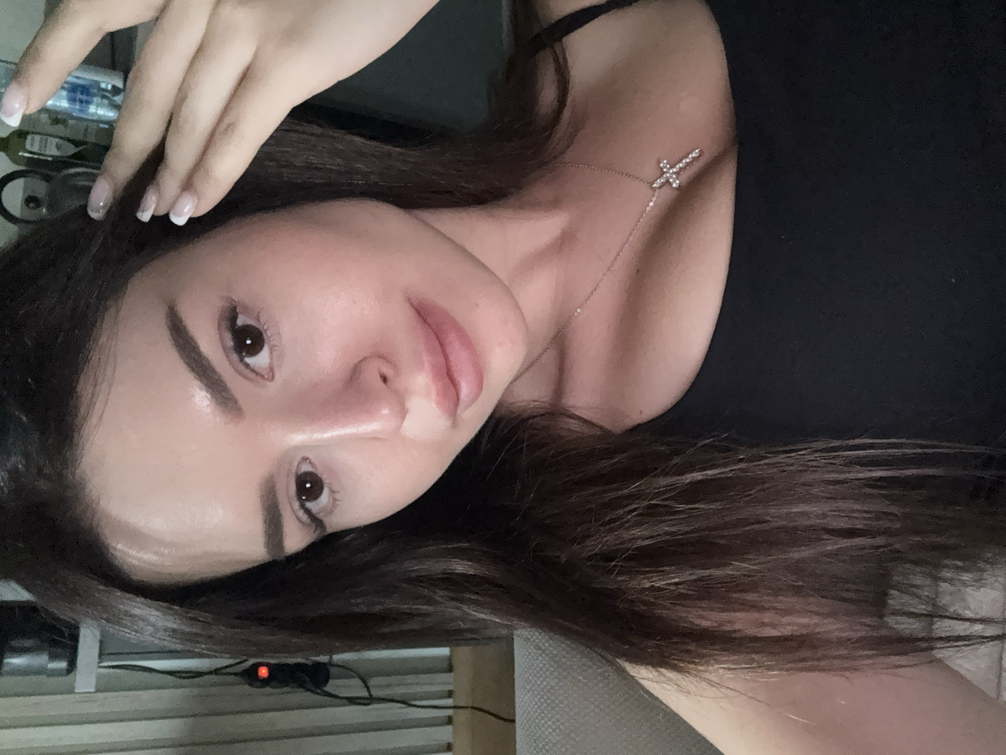 The image is a close-up, indoor, waist-up portrait of a young adult female. She has long, dark brown hair that is styled straight and falls around her shoulders. Her skin is fair, and she has a light application of makeup, with emphasis on her eyes and lips. She is wearing a black, sleeveless top, and a delicate silver necklace with a cross pendant is visible on her chest. Her right hand is raised, touching her hair near her temple. The background is out of focus, but hints of a domestic setting are visible, including what appears to be a shelf with bottles and a patterned curtain or blind on the left side of the frame. There is no visible indication of the city of Prague, Czechia, or any specific urban environment.