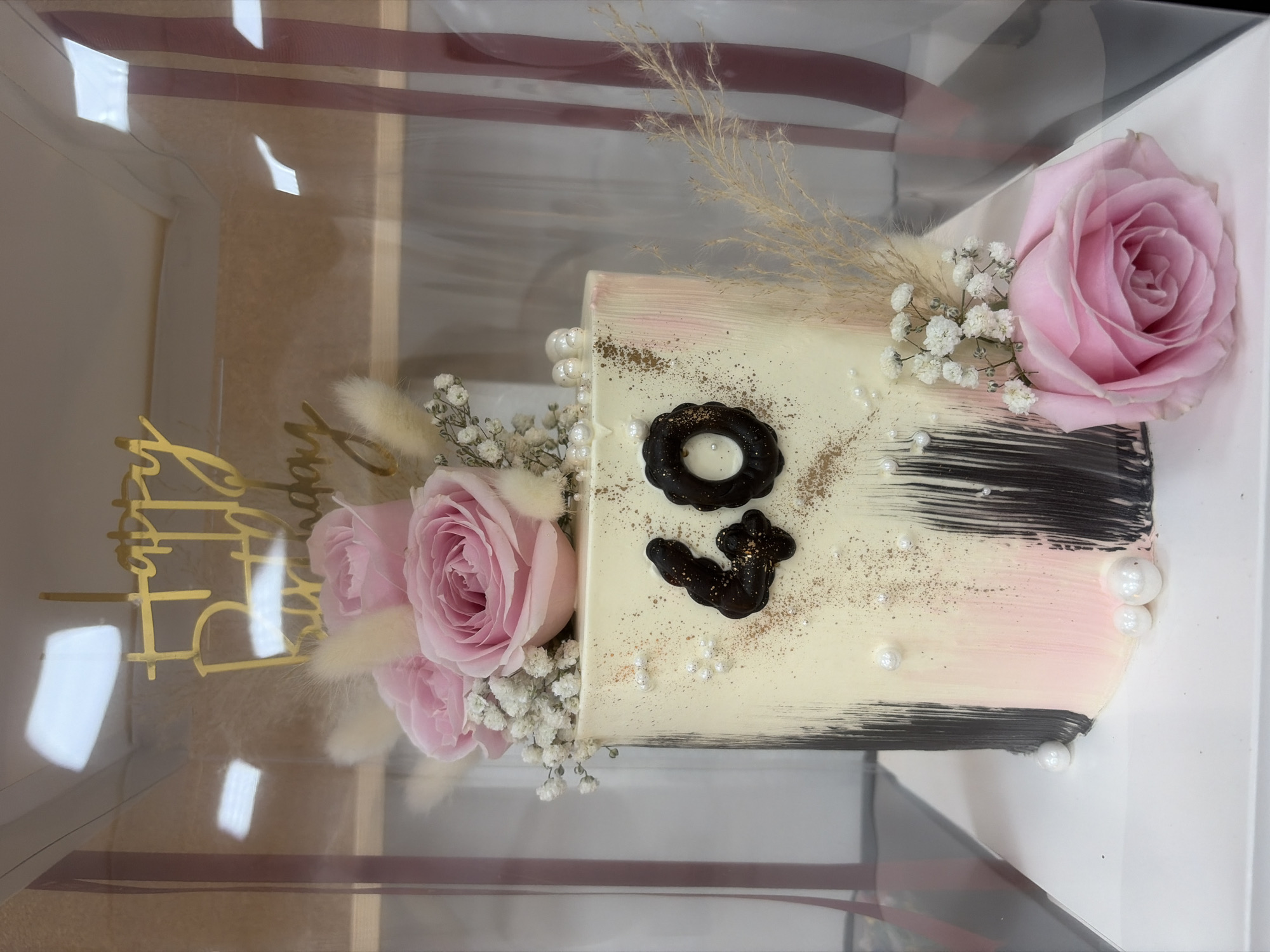 A two-tiered cake, decorated with pink roses and white baby's breath, is displayed in a clear plastic box. The cake features a gold "Happy Birthday" topper and chocolate numbers "40" on the side. The cake itself is white with pink and black brushstroke decorations. The scene appears to be a bakery or a display case. There are no people visible in the image. The background includes reflections of what appears to be lighting fixtures. The location is not specified in the image.