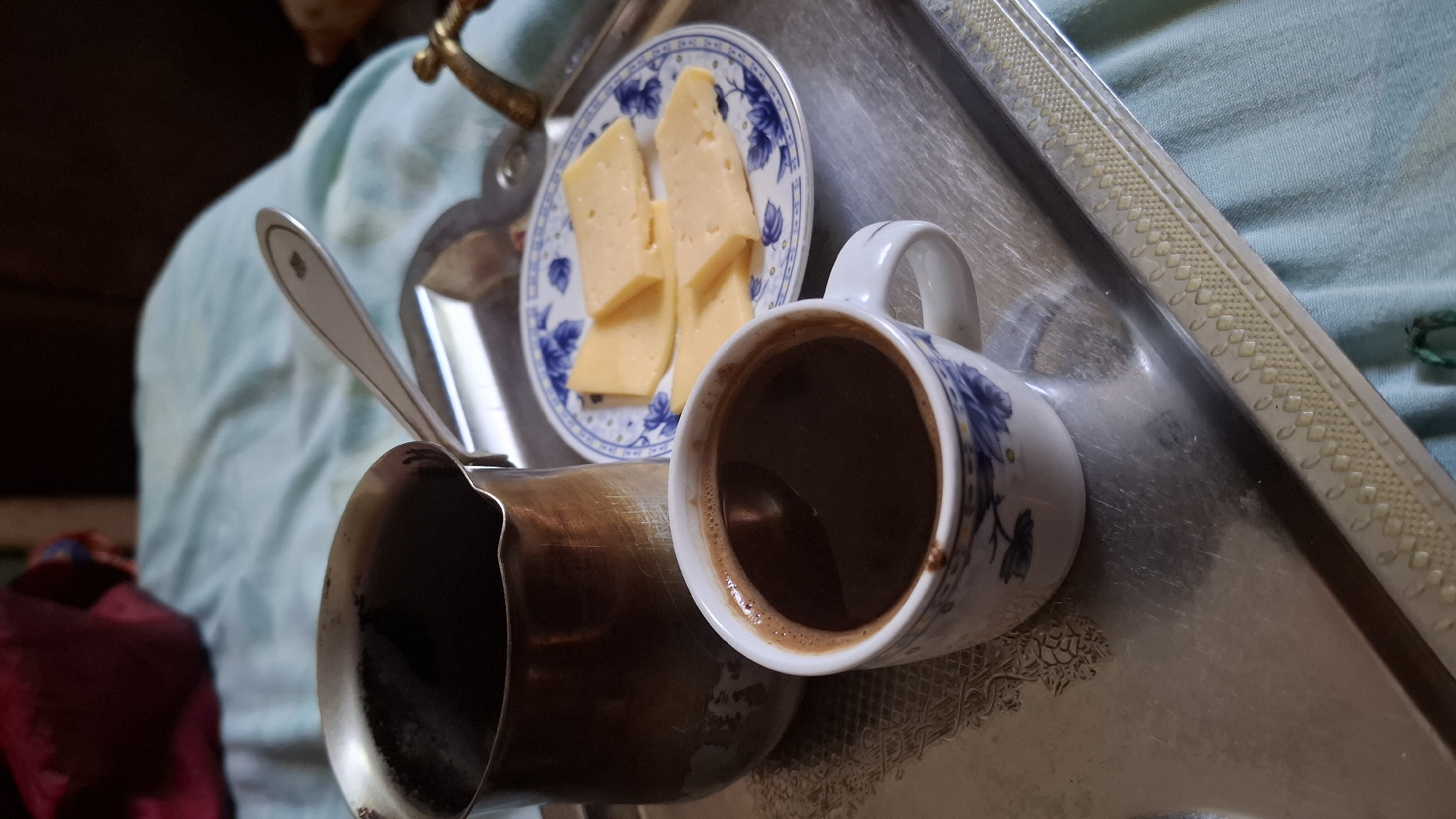 On a tarnished silver tray, a white and blue patterned teacup filled with dark coffee sits next to a copper cezve, a small plate with three slices of yellow cheese, and a silver spoon. The tray rests on a light blue fabric, likely a bedsheet. In the background, a dark red object is visible, possibly clothing, and a section of a brown surface. The overall scene suggests a simple breakfast or snack.