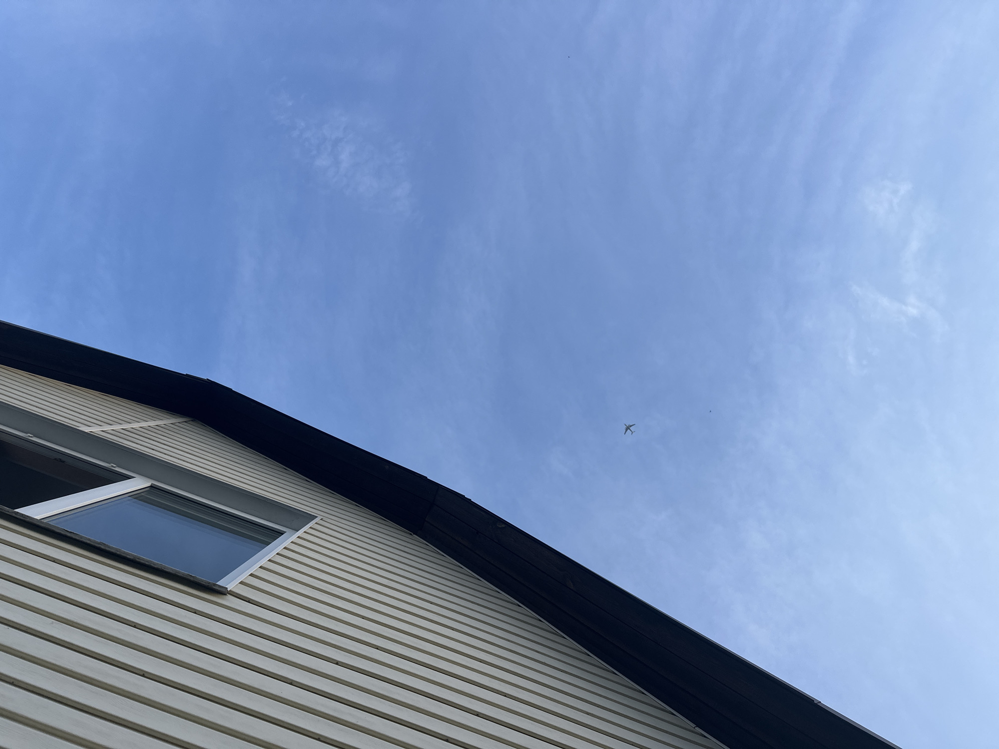 The image shows the exterior of a building with light-colored horizontal siding and a black roofline. A window is visible on the left side of the building. The majority of the image is filled with a blue sky containing wispy clouds. A small airplane is visible in the sky. It's impossible to confirm the location as Nizhny Novgorod, Russia based on the image alone. There are no visible people, specific objects, or actions to further characterize the scene.