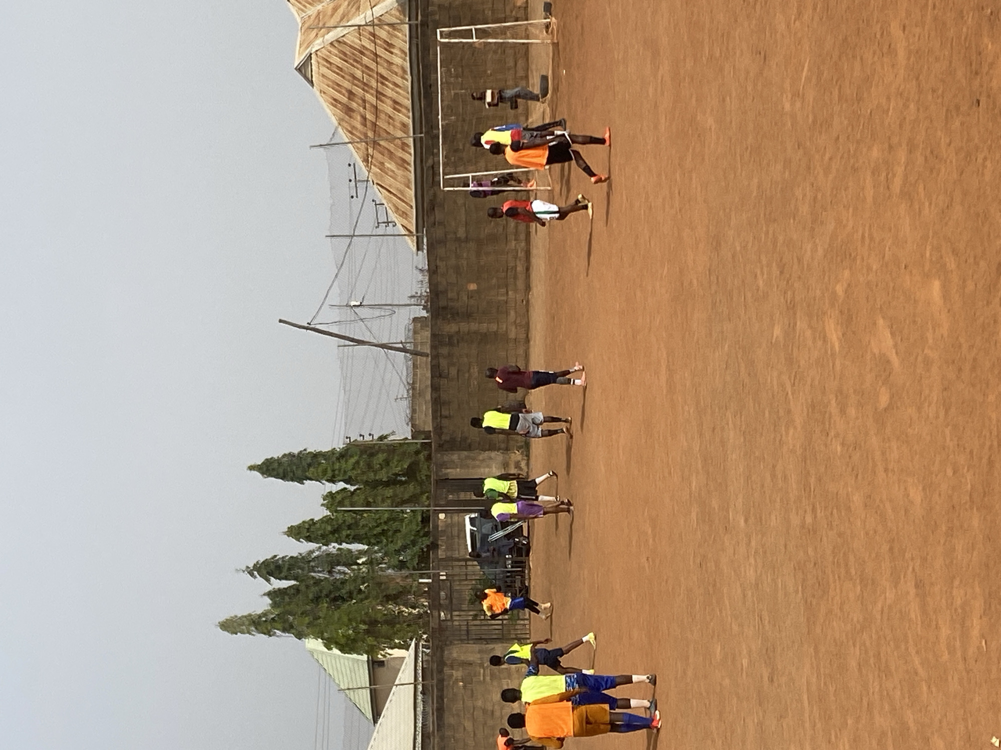 The image captures an outdoor scene featuring a group of individuals engaged in physical activity on a dirt field, likely in the city of Jos, Nigeria. A row of young men are performing push-ups, dressed in a variety of athletic wear including shorts and t-shirts in colors like yellow, blue, orange, and burgundy. In the background, a soccer goalpost is visible, along with a low brick wall and a building with a corrugated metal roof. Trees line the side of the field. Overhead power lines are visible against a hazy sky.