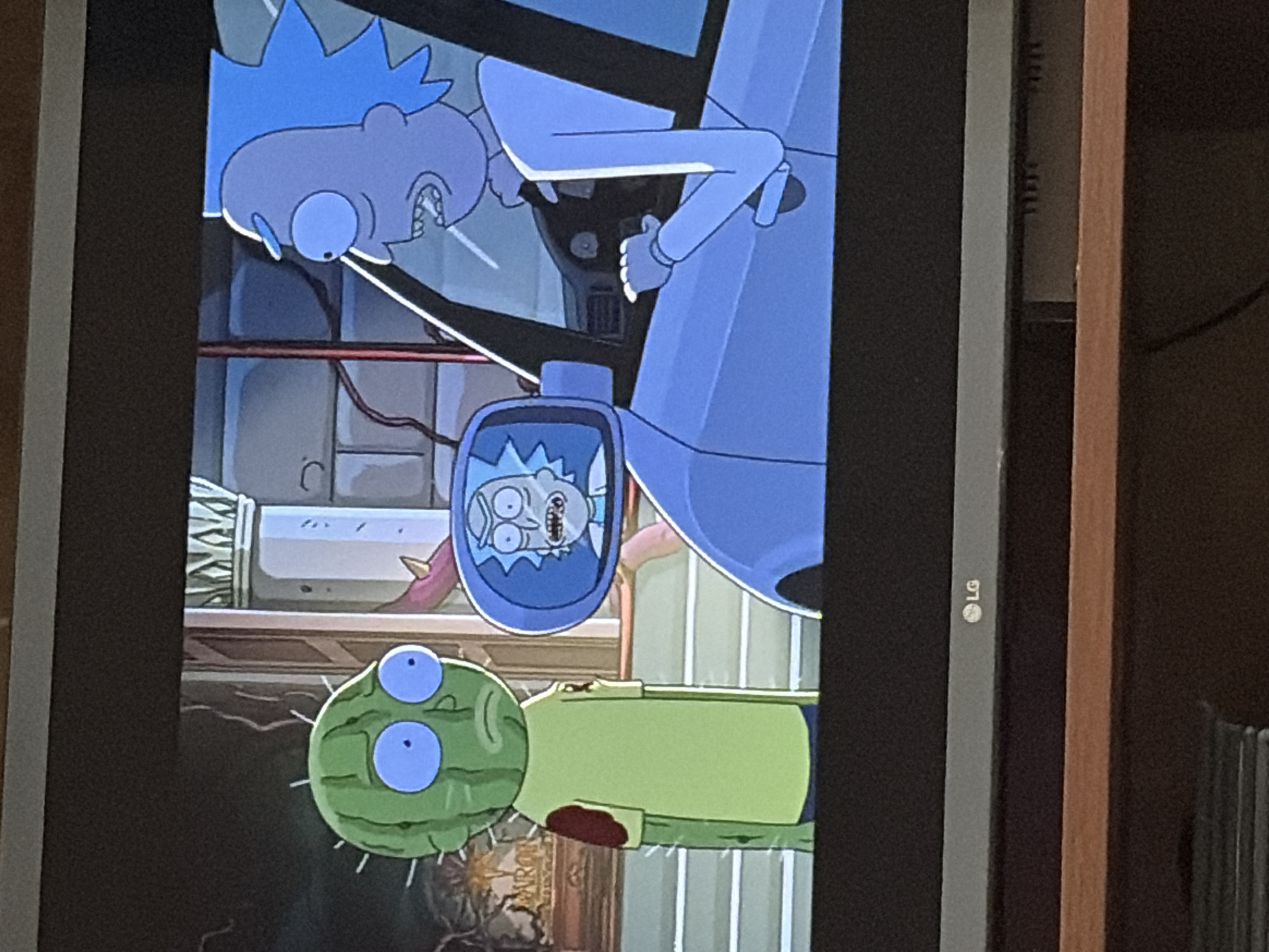 In an animated scene, a cactus-like creature with large blue eyes and a yellow shirt stands to the left. To the right, a blue-haired, humanoid character, resembling Rick Sanchez, is driving a light blue vehicle. A side mirror reflects a second image of the same blue-haired character, appearing distressed. The background suggests an indoor environment with architectural elements and ornate decorations.