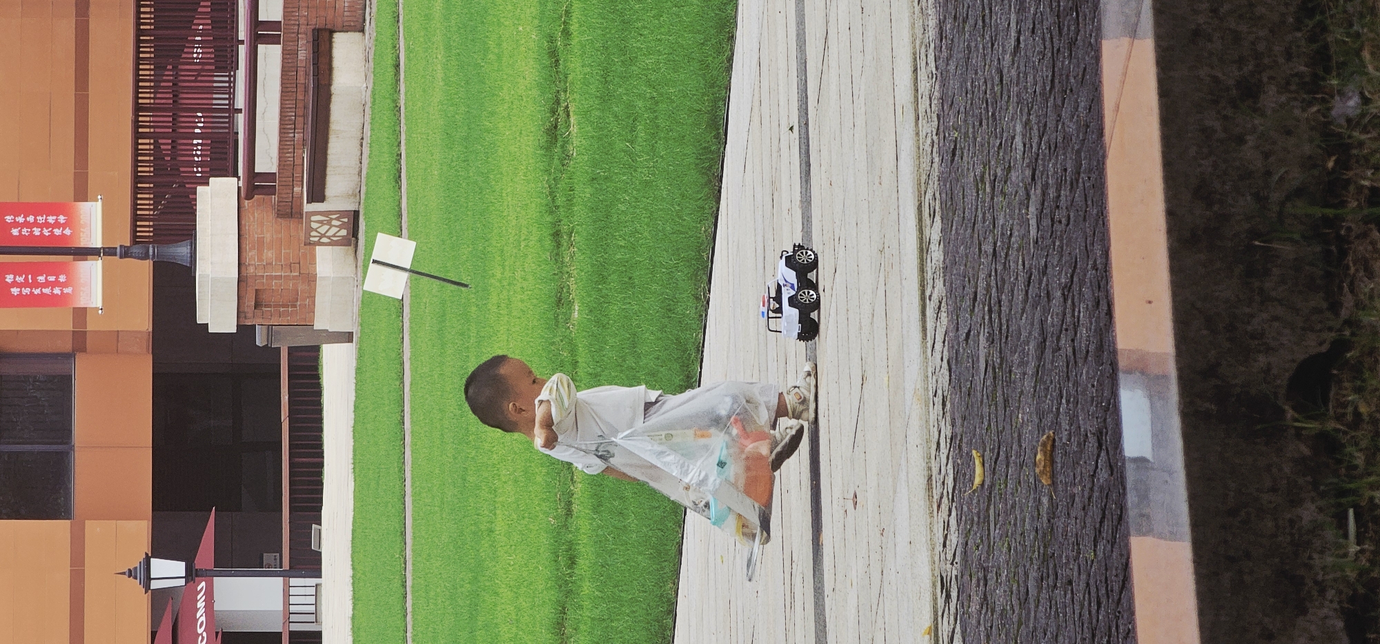 The image shows a young child carrying a plastic bag and walking on a paved path in a green space. The location is in Yuzhong District, China, indicated by Chinese banners with the name “COMU” displayed in Latin script. There is a toy police car on the path near the child. The area appears to be a public space with grass and building facades.