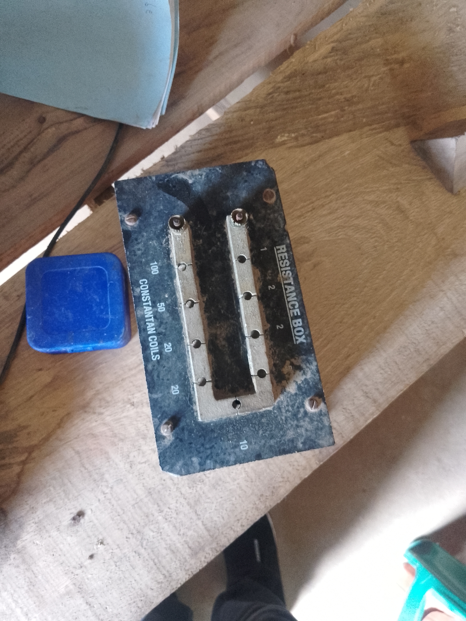 The image shows a resistance box with numerical labels and "RESISTANCE BOX" and "CONSTANTAN COILS" printed on it, placed on a wooden surface. A blue plastic lid is also visible next to the resistance box. The background consists of wooden planks and a light blue folder. There are no people or identifiable landmarks present in the image. The scene does not provide any information to determine its location in Serti, Nigeria.