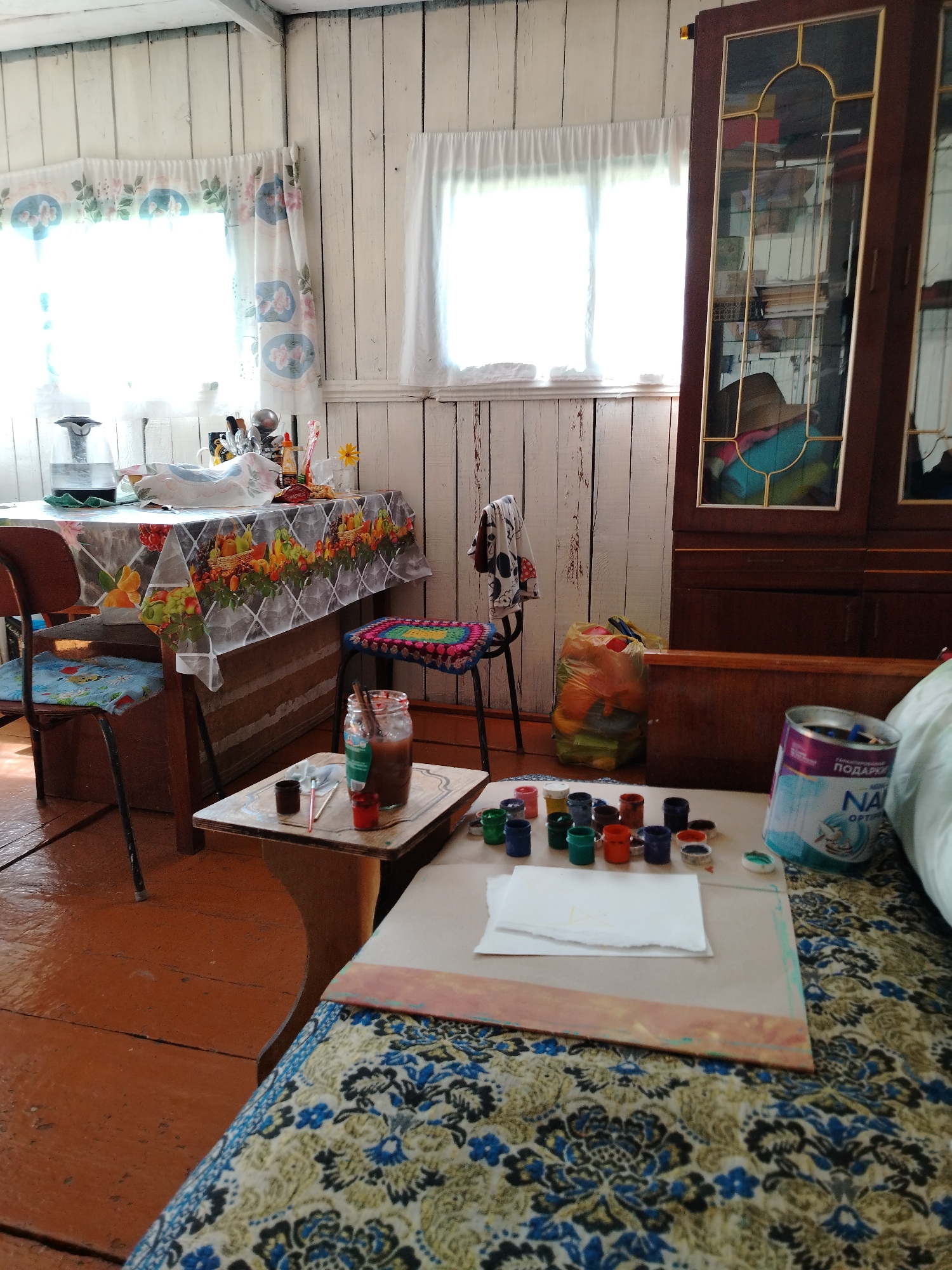 The image depicts the interior of a room, likely a cottage or a rural dwelling in Iodro, Russia. The walls are paneled with light-colored wood, and the ceiling beams are also visible. Natural light streams in from two windows covered by floral patterned curtains on the left and sheer white curtains on the right.
In the foreground, a patterned blanket or tablecloth is spread over a surface, possibly a couch or a low table. On this surface, a collection of small paint pots is arranged, with various colors visible. A sheet of white paper is placed in front of the paint pots. To the right, a can of "NA OPTI" product, possibly a cleaning or laundry detergent, is standing upright.
A small wooden table sits to the left of the main foreground surface. On this table, a glass jar with a brown substance and a brush or stirring stick inside is placed. Next to it are small containers of paint and a red object. A chair with a blue cushion is positioned by a larger table.
This larger table is covered with a patterned tablecloth featuring images of fruit and flowers. On the table, various items are arranged, including what appears to be a kettle and some containers. A wooden cabinet with glass doors stands against the wall on the right side of the image, its shelves filled with items. A plastic bag filled with colorful items, possibly toys or fabric scraps, is leaning against the cabinet. A stool with a colorful crocheted seat is placed near the larger table. The floor is made of wooden planks with a reddish-brown finish. The overall impression is of a lived-in space, possibly used for artistic or craft activities.