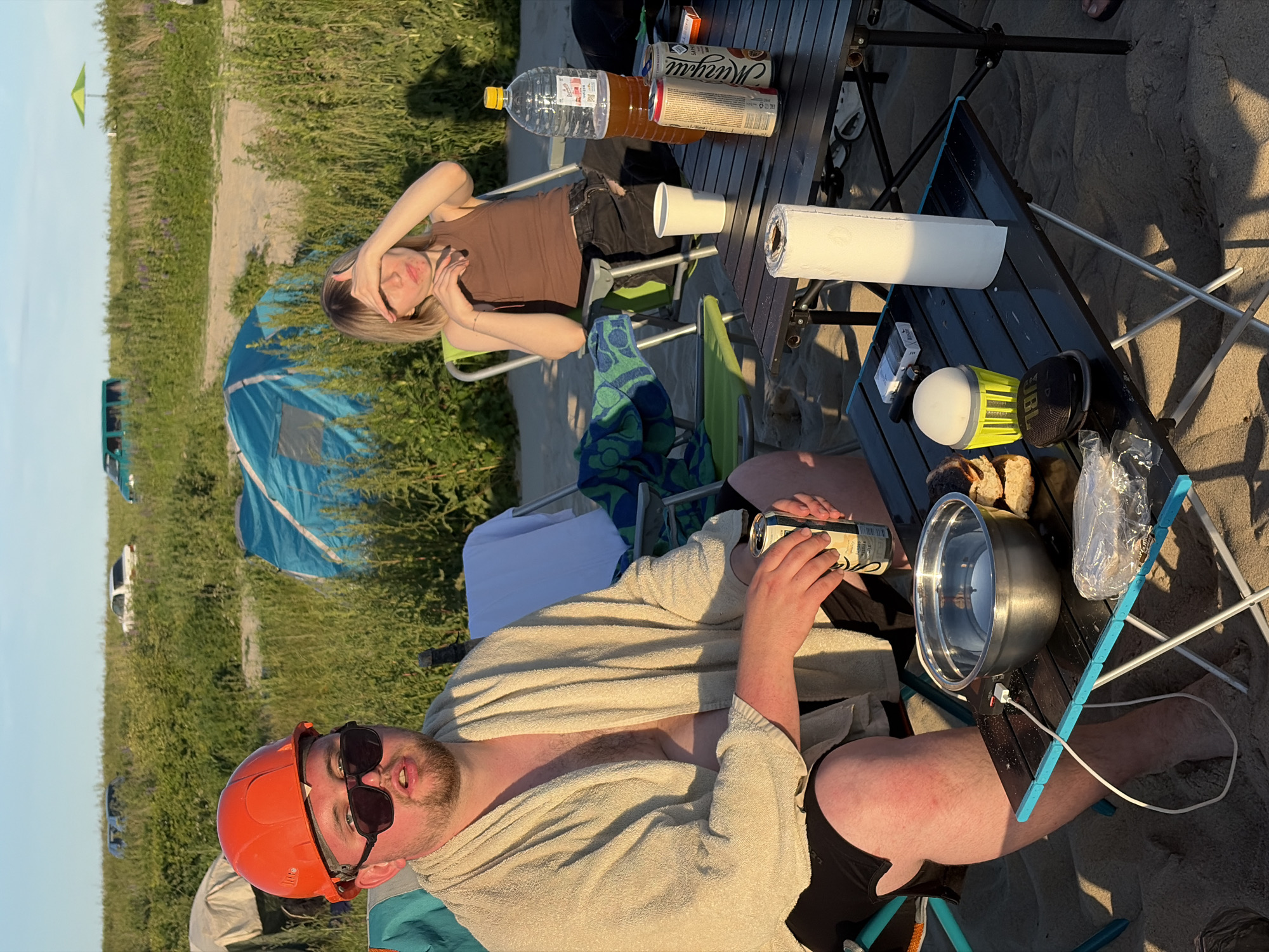 The image captures a sunny outdoor scene on a sandy beach, likely in Povarovka, Russia, given the context. In the foreground, a man wearing an orange hard hat and sunglasses is seated in a lawn chair. He is draped in a light-colored towel and holding a can of soda. To his right, a black folding table holds several items, including a metal bowl, bread, a roll of paper towels, a small light, a pack of cigarettes, and a portable speaker. Further back, a young woman with blonde hair is seated in a green lawn chair, with her hand shielding her eyes from the sun. Next to her, a large plastic bottle of amber liquid and two cans are visible on a table. In the background, a blue tent is set up, along with other camping gear and what appear to be parked cars and a green umbrella. The environment is characterized by tall grasses and a sandy beach under a clear sky with warm, late afternoon sunlight casting long shadows.