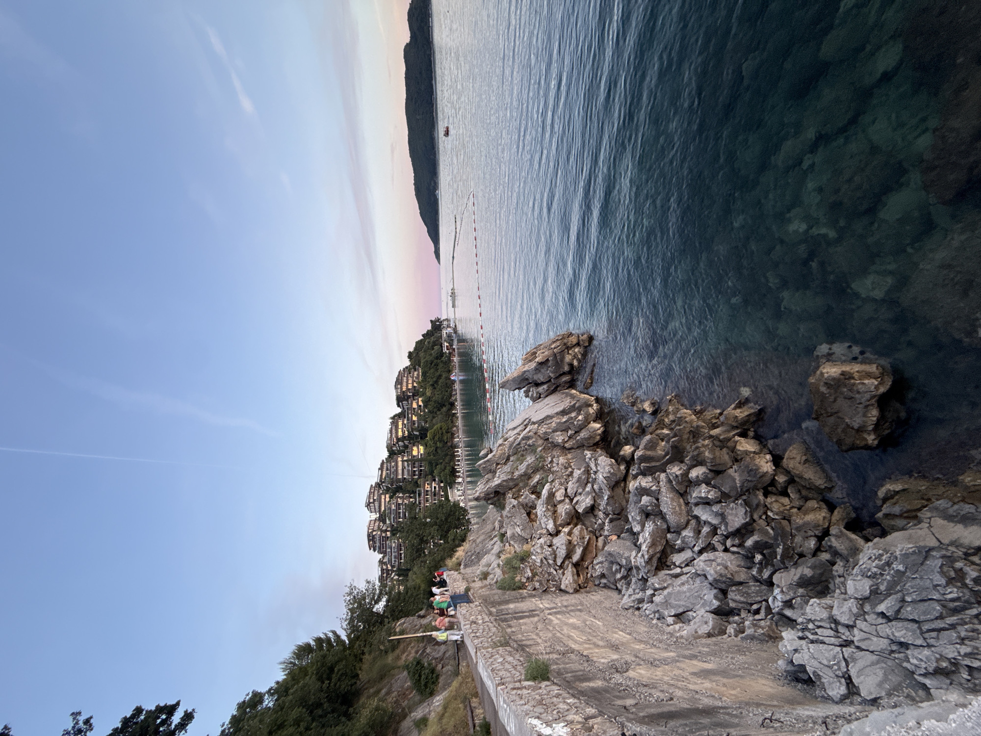 The image captures a view of the Adriatic Sea coastline near Budva, Montenegro, likely during the golden hour or twilight. In the foreground, a stone-walled promenade runs along a rocky shore. Several people are visible walking on the promenade, some casually observing the sea. The sea itself is a deep blue-green, with clear visibility of the rocky seabed in shallower areas. A red and white buoy line stretches across the water in the middle ground, indicating a designated swimming area or boundary. Further in the distance, a chain of modern, multi-story buildings is nestled into the green, hilly terrain that slopes down to the water. These buildings appear to be hotels or apartments, characteristic of resort architecture. On the right side of the frame, a small, dark island or peninsula juts out from the horizon, its silhouette softened by the atmospheric light. The sky above transitions from a clear, pale blue to a soft pink and lavender gradient near the horizon, suggesting the sun is setting or has recently set. The overall scene conveys a sense of tranquility and coastal beauty.