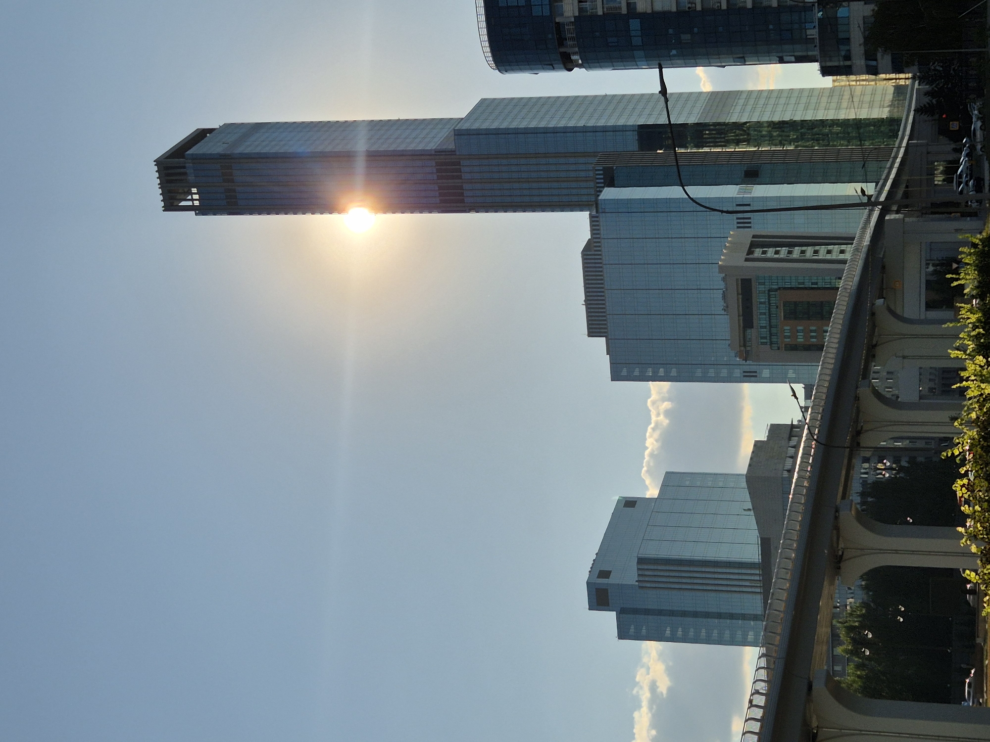 The photograph captures a daytime, exterior view of a modern cityscape in Astana, Kazakhstan, dominated by contemporary architecture and a clear sky. The sun is prominently featured, positioned high in the sky and partially obscured by a tall, slender skyscraper, creating lens flare that radiates outwards. This skyscraper, a central element, is characterized by its glass facade and a distinct, open-topped structure at its summit.
In the foreground, an elevated structure, likely a roadway or pedestrian bridge, spans across the scene, supported by arched pillars. This structure is flanked by lush green foliage and trees, suggesting the presence of urban green spaces.
Several other modern buildings with glass exteriors are visible, including a wider, shorter building with a distinctive angular roofline to the left of the main skyscraper, and a curved, dark-glass building to its right. The reflections on the glass facades of these buildings mirror the sky and surrounding structures.
Below the elevated walkway, a street level scene is partially visible, with parked cars and streetlights. The overall impression is one of urban development and a bustling metropolitan environment under a bright, sunny sky, characteristic of Astana. There are no discernible people in the image. The lighting suggests it is late afternoon or early morning, given the angle of the sun and the warm tones.