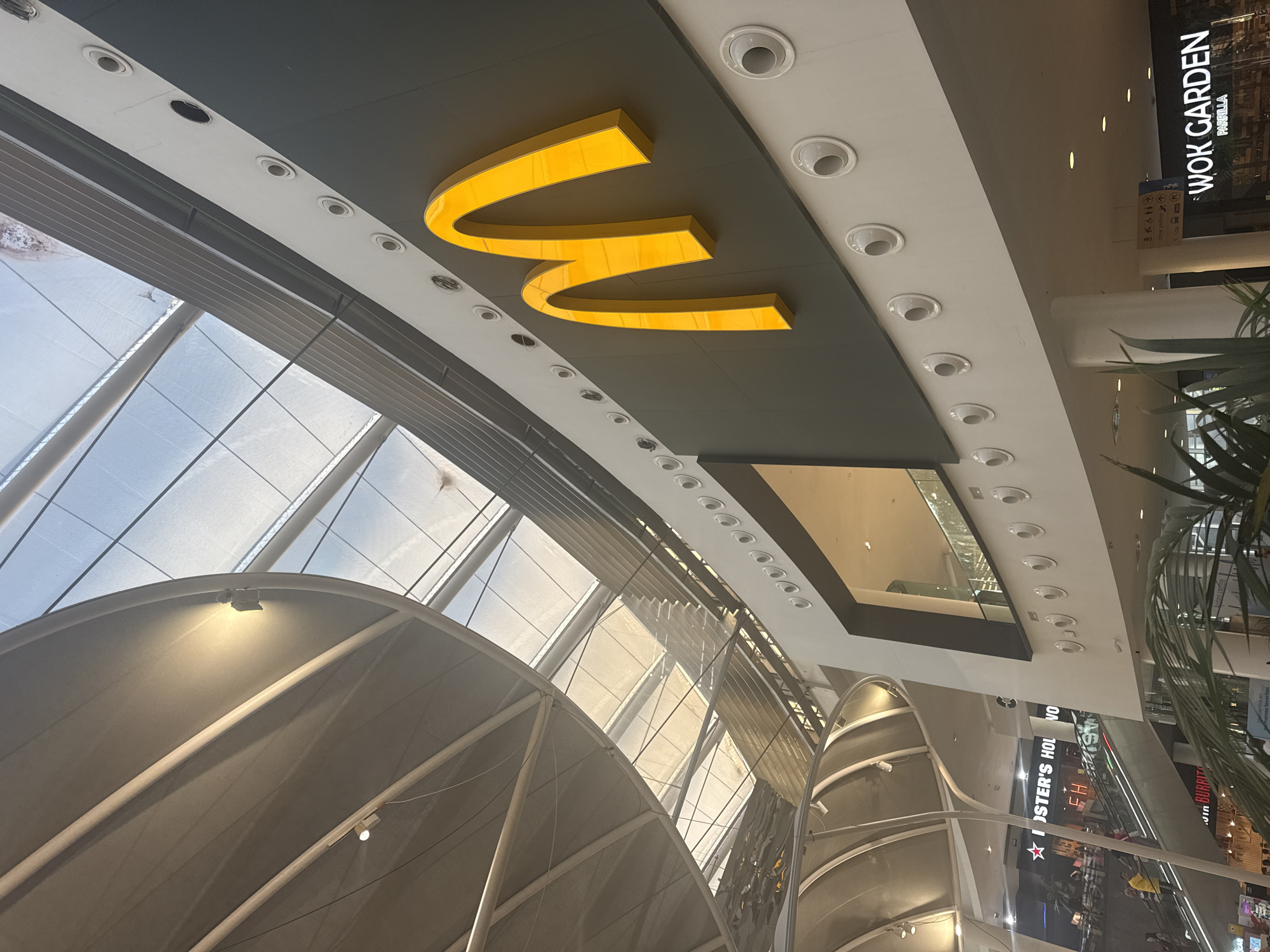 The photograph captures an interior view of a modern shopping mall with a prominent McDonald's sign. The McDonald's logo, the iconic golden arches, is illuminated and mounted on a dark grey panel on the upper right side of the image. Below this panel, on a white surface, are several circular vents or light fixtures. The ceiling of the mall features a complex architectural design with large, curved translucent panels that allow natural light to filter through, creating a bright, airy atmosphere. Metal beams and supports are visible within the ceiling structure.
In the background, on the left side of the image, a large escaltor is visible with several people on it, suggesting the mall is open and active. Signs for other businesses, such as "FOSTER'S HOLL" and "WOK GARDEN PARRILLA," are also partially visible, indicating a variety of retail and dining options. The overall environment appears clean, modern, and well-lit. The style of the architecture, particularly the ceiling and the facade of the McDonald's outlet, suggests a contemporary shopping center. Based on the presence of a McDonald's and the overall design, this scene is likely from a shopping mall in Madrid, Spain.