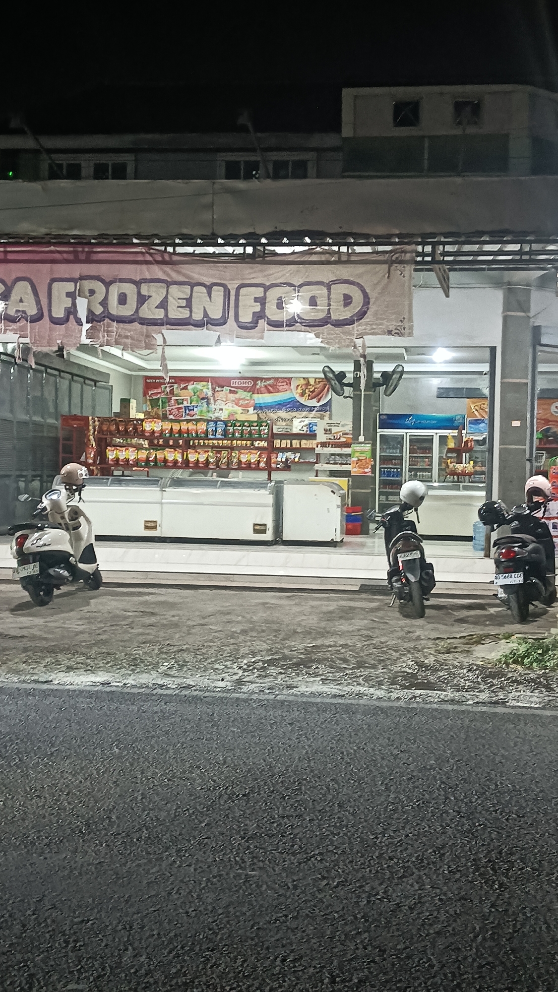 A low-angle, outdoor, medium shot captures a frozen food store at night on a street in Sragen, Indonesia. The store's facade is illuminated, revealing a large banner overhead that reads "A FROZEN FOOD." Inside, shelves are stocked with various packaged goods, and several large, white, horizontal freezers are visible in the foreground. A light-colored scooter with a helmet on its seat is parked on the left, and two darker-colored scooters with helmets are parked in front of the store on the right. The street in front of the store is paved and appears wet. The background shows a two-story building with windows and structural elements characteristic of Indonesian architecture. The overall lighting suggests it is nighttime, with artificial lights from the store and street illuminating the scene.