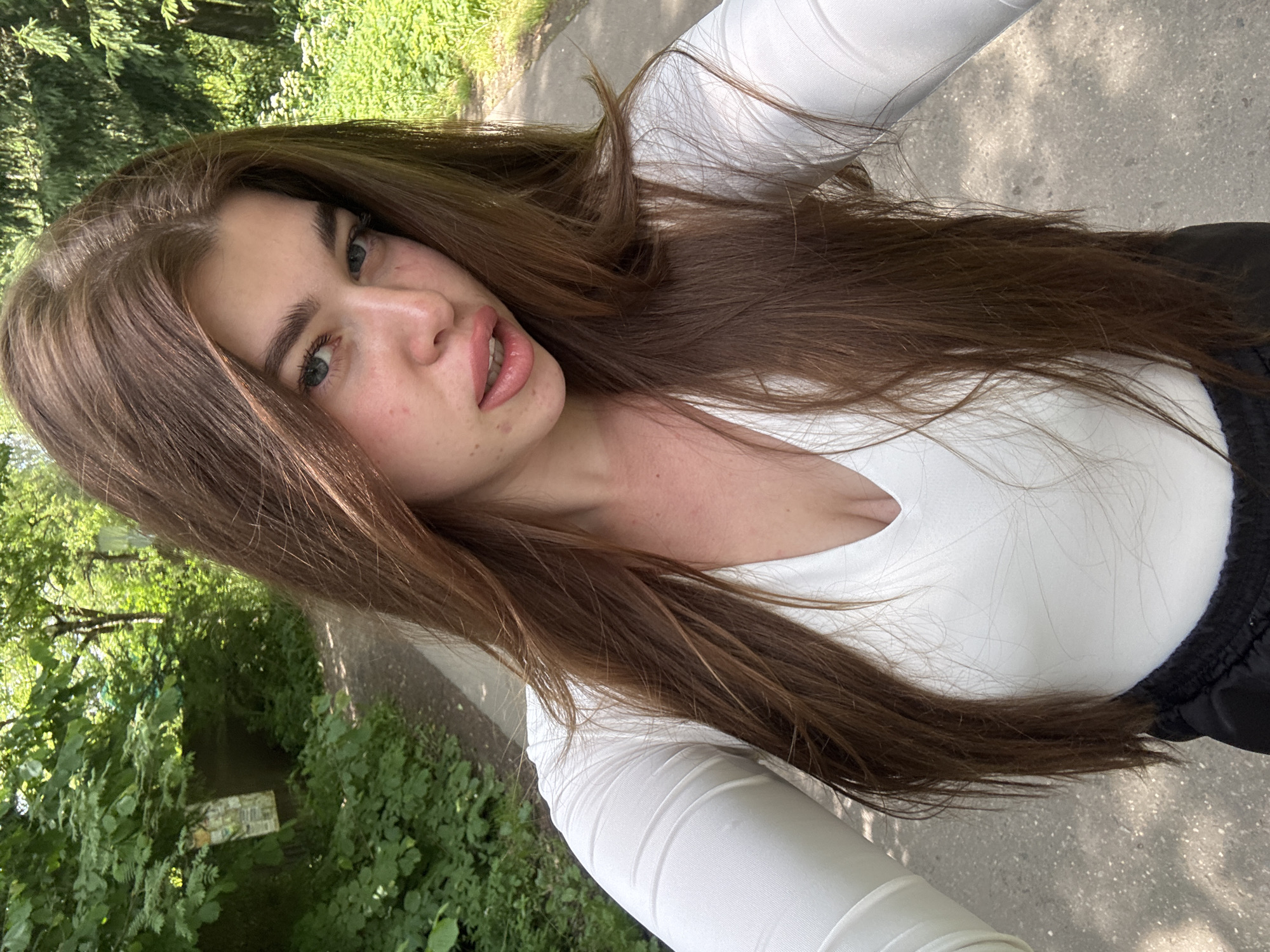 This is a selfie of a young woman with long, brown hair, taken outdoors during the daytime. She is wearing a white long-sleeved shirt and black athletic pants. Her hair is dark brown and falls over her shoulders and chest. She has fair skin with some freckles visible on her face and neck. Her eyes are light blue-green, and she is looking towards the camera with her mouth slightly open, revealing her teeth. The background suggests a natural setting, with lush green trees and foliage on the left side of the image. A paved path runs behind her, with dappled sunlight indicating a sunny day. The overall mood is casual and relaxed. There is no visible text in the image. The location context is Мещёры, Russia.