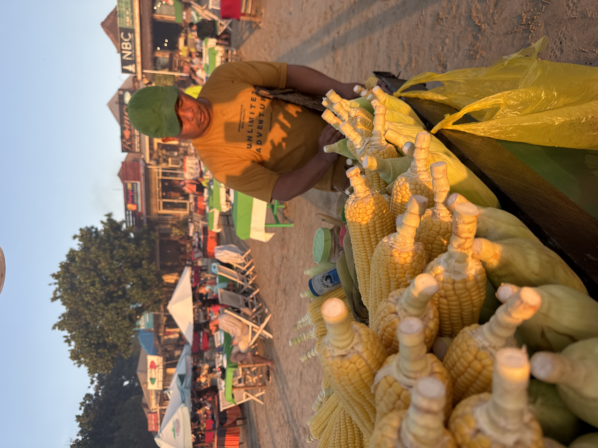 The image shows a street vendor in Kuta Selatan, Indonesia, selling corn on the cob. The vendor, wearing a green cap and mustard-colored t-shirt, is positioned behind a cart laden with corn. Yellow plastic bags are also visible on the cart. In the background, there is a beach scene with various buildings, chairs, and umbrellas. The sky is clear and appears to be late afternoon or early evening.