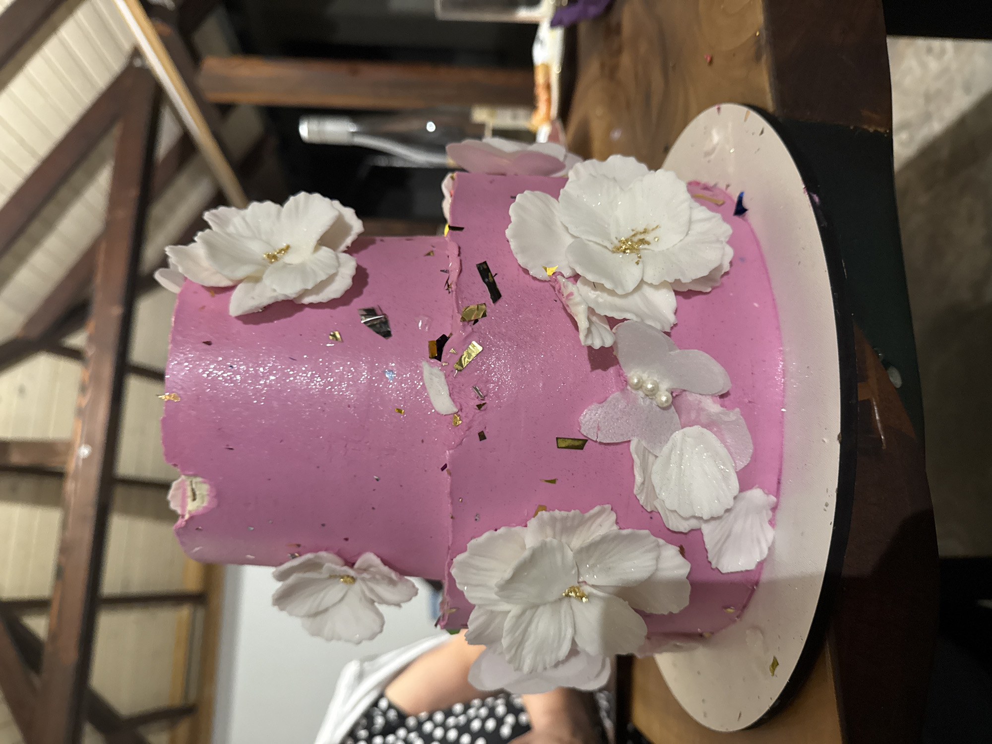 The image shows a two-tiered pink cake decorated with white edible flowers and gold flakes. The cake is placed on a wooden table, likely in an outdoor setting given the visible wooden beams and ceiling structure in the background. A person is partially visible on the left side of the image, wearing a black and white polka dot garment. In the background, a bottle and a glass are also present on the table. The scene appears to be a celebration or event, possibly in Verejeni, Moldova.