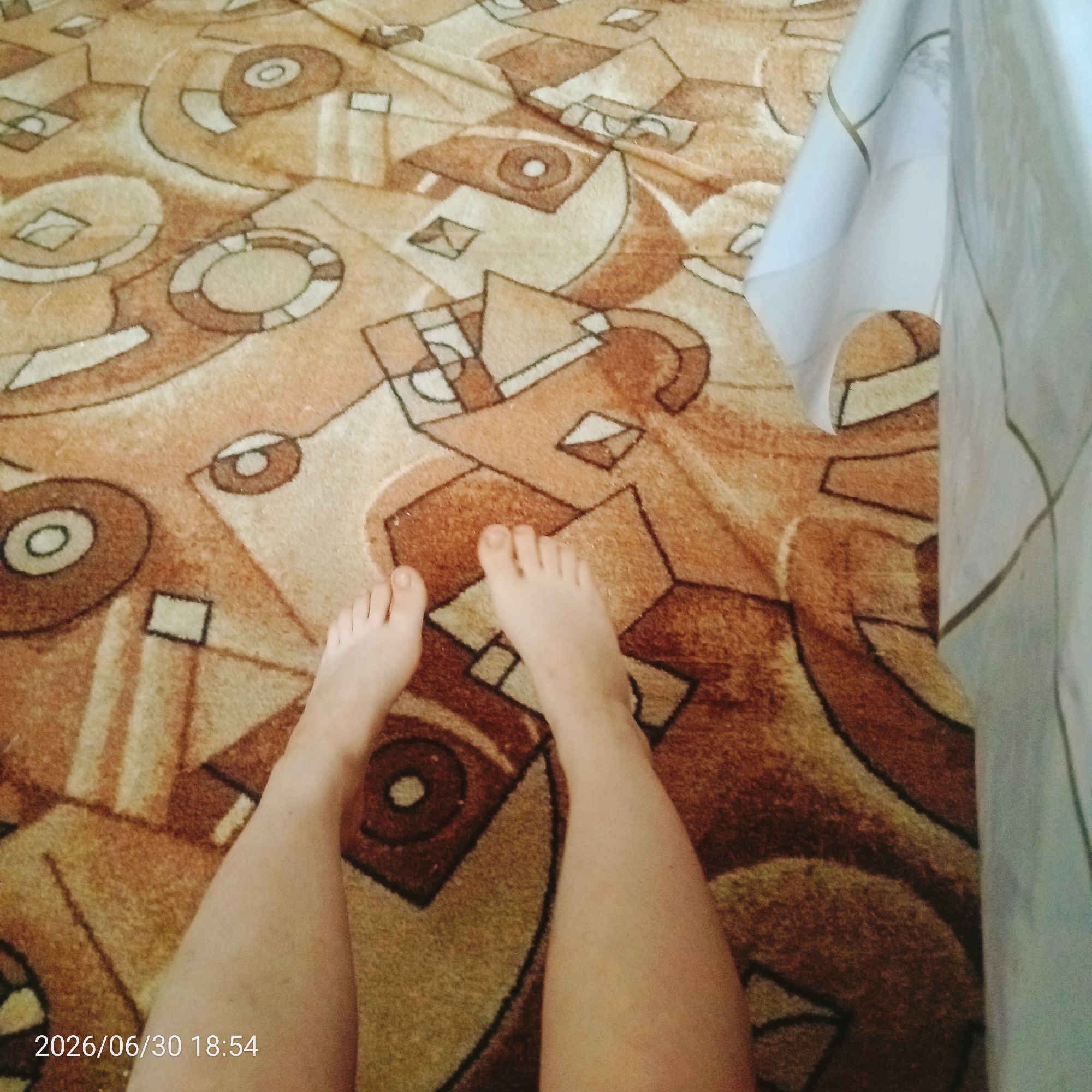This media file shows a close-up of a person's legs and feet resting on a patterned carpet. The carpet has a warm color palette of browns, tans, and beige, with an abstract geometric design. The person's legs and feet are bare. The lower right corner of the image displays a timestamp: "2026/06/30 18:54", indicating the date and time the photo was taken. A light-colored fabric, possibly a curtain or tablecloth, is visible on the right side of the frame. The scene appears to be indoors, likely in a home setting, given the carpet. The lighting suggests it is either late afternoon or early evening. There are no specific emotions conveyed by the image, and the focus is on the textures and patterns of the carpet and the person's legs.