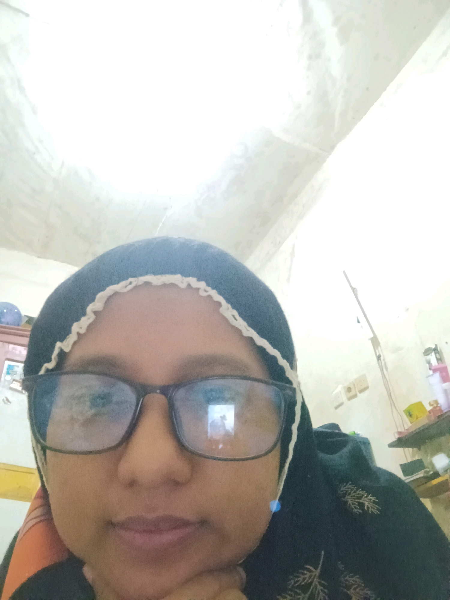 A close-up, eye-level, medium shot captures a person wearing a dark hijab with a white trim and dark-rimmed glasses. The individual is resting their chin on their hand, looking towards the camera. The background is dimly lit with a textured, light-colored ceiling and a white wall. To the right, there are some shelves with various items, including bottles and what appears to be a laptop. The overall setting suggests an indoor environment, possibly a room in SARANGAN, Indonesia.