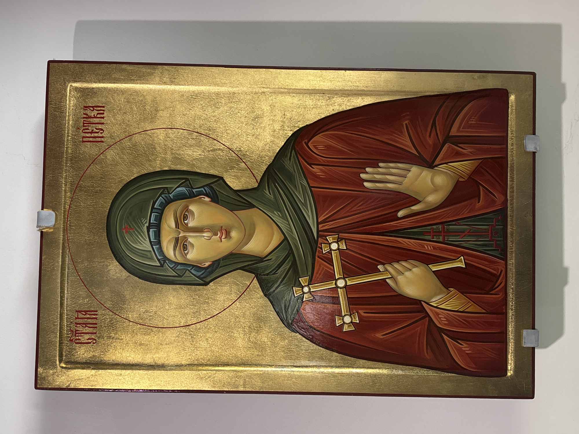 The image presents a vertically oriented Eastern Orthodox icon of Saint Petka, displayed against a plain white wall in Belchin, Bulgaria. The icon features a gold leaf background with a textured finish, bordered by a reddish-brown frame and an inner raised gold border.
The central figure is a youthful female saint, identified by the Cyrillic inscription "СТЛА ПЕТКА" (Saint Petka) in red lettering at the top left and right corners. She wears a dark green maphorion, which covers her head and shoulders, revealing only her face. Beneath the maphorion, a lighter blue-grey undergarment is visible, and a small red cross adorns the fabric over her forehead. Her main outer garment is a deep red robe, characterized by stylized folds, with gold bands visible on the sleeves at her wrists. A thin red halo encircles her head.
The saint's proper left hand is raised in a gesture with the palm open and fingers slightly spread. Her proper right hand holds a golden martyr's cross, which features quatrefoil-shaped terminals, each with a small circular boss at its center.
The icon is supported at the bottom by two small, light-grey rectangular blocks. A similar block is visible at the top center, likely serving as a hanger. The lighting is even, producing subtle shadows on the white wall behind the icon and its supports.