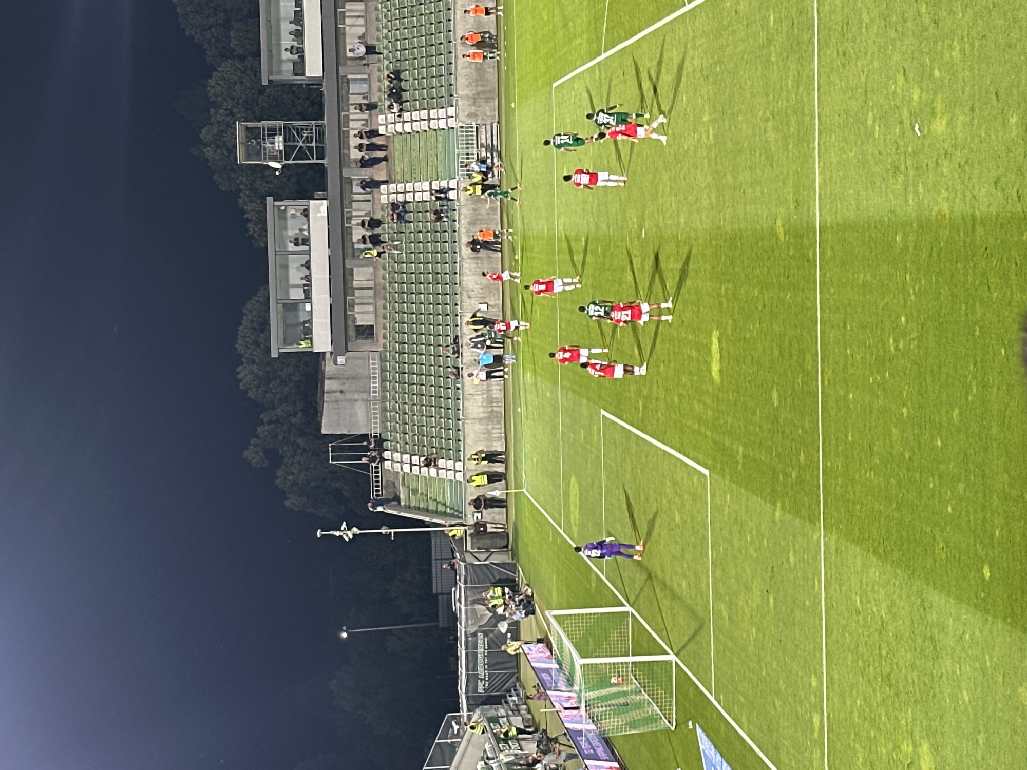 The image depicts a soccer match at night on a grass field. The stadium lights illuminate the field, casting shadows from the players. Spectators are visible in the tiered green seating areas, with some standing in the open sections. Several individuals in yellow vests, likely security or staff, are positioned near the sidelines and entranceways. The overall atmosphere suggests an active sporting event. The stadium is located in Razgrad, Bulgaria.