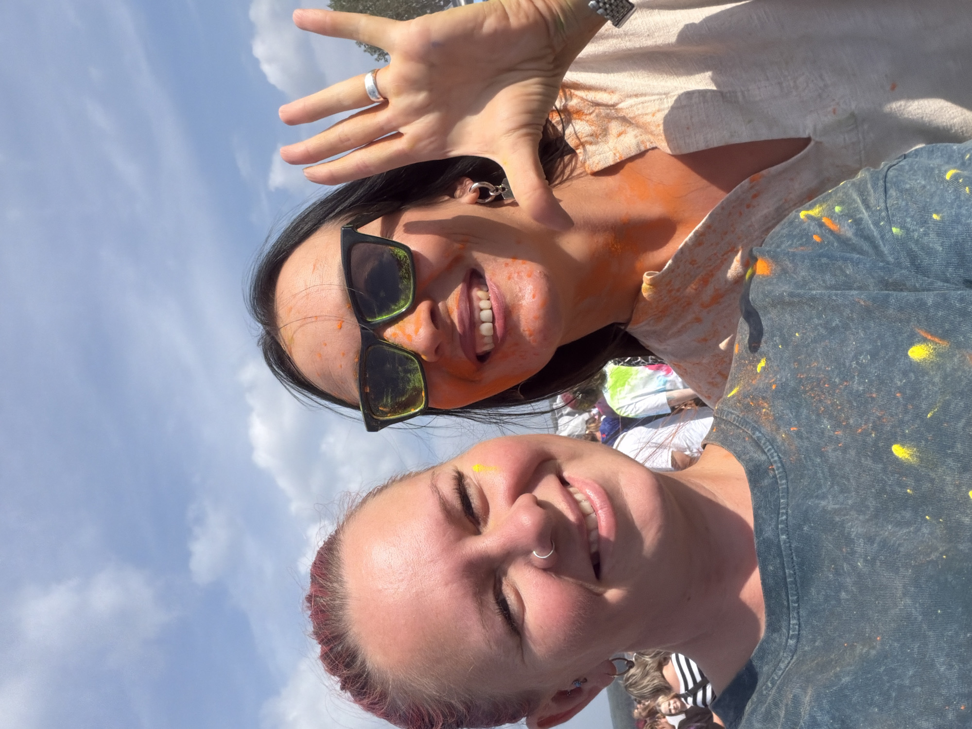 Two women are posing for a selfie outdoors on a sunny day, with a blue sky and some clouds visible in the background. Both women have been participating in a color festival, as evidenced by the colorful powder splattered on their faces, necks, and clothing. The woman on the left has red hair pulled back, a nose ring, and is smiling. The woman on the right is wearing sunglasses and has her hand raised, showing off a ring on her finger. She is also smiling, and her face and neck are covered in orange powder. In the background, other people are visible, also appearing to be covered in colored powder, suggesting a public event. The location is identified as Chaikovsky, Russia.