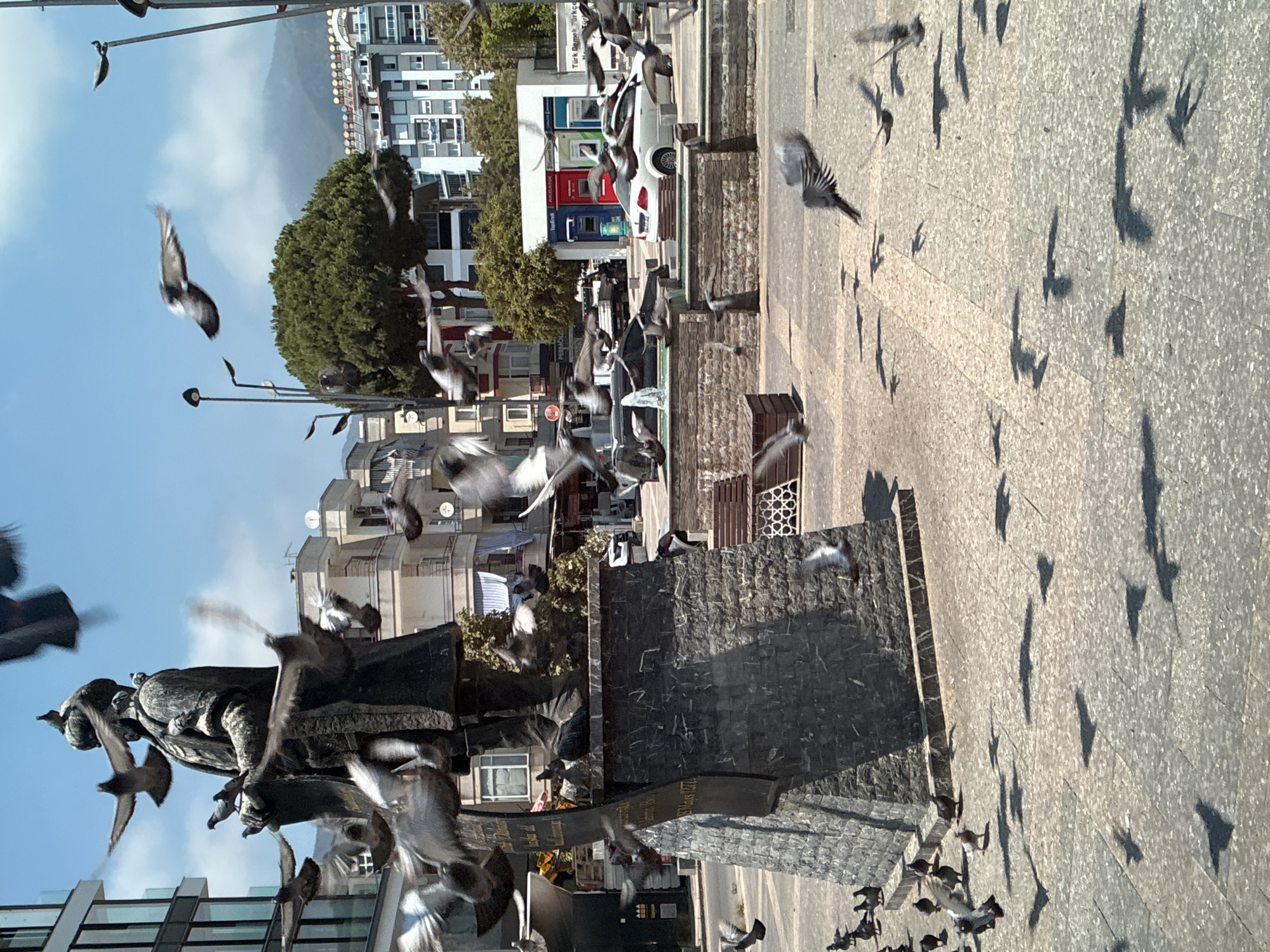 A statue of a man in traditional attire stands prominently in a paved square, surrounded by numerous pigeons in mid-flight. The statue is dark in color and appears to be made of stone or bronze, with a crown on its head. The pigeons are captured in various stages of flight, with some wings blurred due to motion. In the background, buildings of varying heights and architectural styles line a street, suggesting an urban environment. A white car is parked near a building with blue and red facades, possibly a bank or an ATM service area. Trees and streetlights are also visible, contributing to the city scene. The sky is clear and blue with some scattered clouds. The overall impression is a lively public space in Alanya, Turkey, marked by the presence of a historical statue and a flock of birds.
