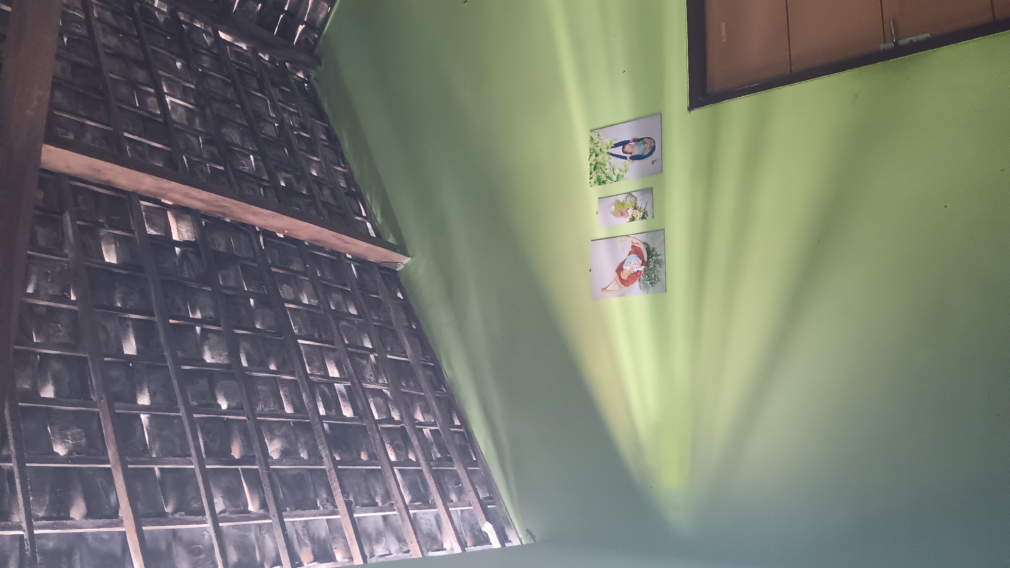 The image depicts an interior view of a room with a light green wall. The ceiling is made of exposed wooden beams and dark clay tiles, with sunlight filtering through the gaps. Three small framed pictures are hung on the wall: two larger ones showing babies in baskets or hammocks, and one smaller one in between them with an unclear subject. To the right of the pictures, part of a brown wooden door is visible, with a silver handle. The lighting in the room is dynamic, with strong shafts of sunlight creating patterns on the wall. The scene appears to be from a residential setting, possibly in Sonobekel, Indonesia, given the architectural style of the roof and the general ambiance. There are no people visible in the image.