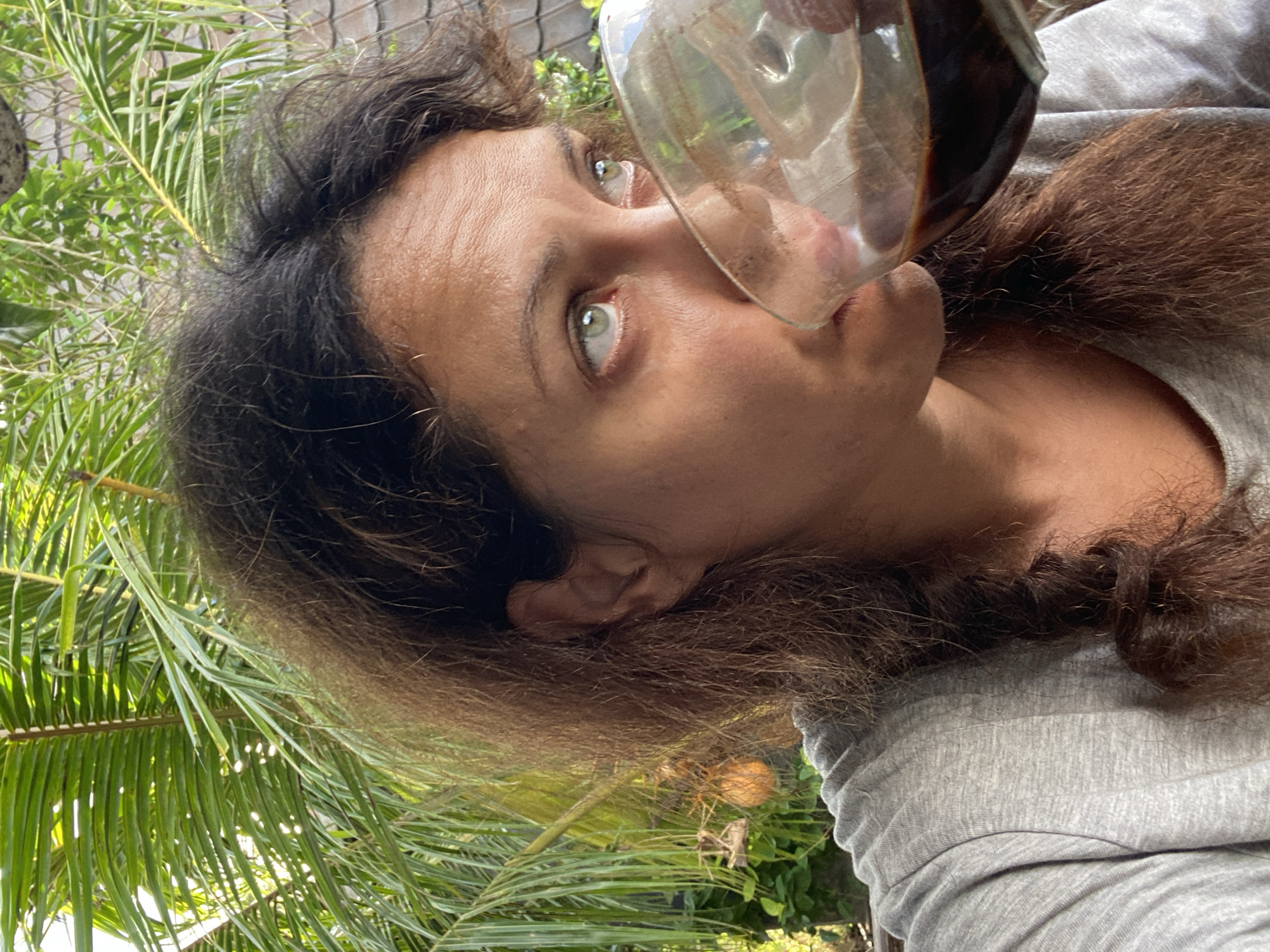 A medium shot captures a woman with long, dark brown hair drinking from a glass. She is wearing a gray t-shirt and her hair is partially covering her face and neck. The woman's eyes are green and she is looking to her right. The background features lush green palm fronds and some vegetation, suggesting an outdoor setting, possibly a tropical location. A woven roof structure is visible in the upper right portion of the frame. The overall lighting suggests daylight.