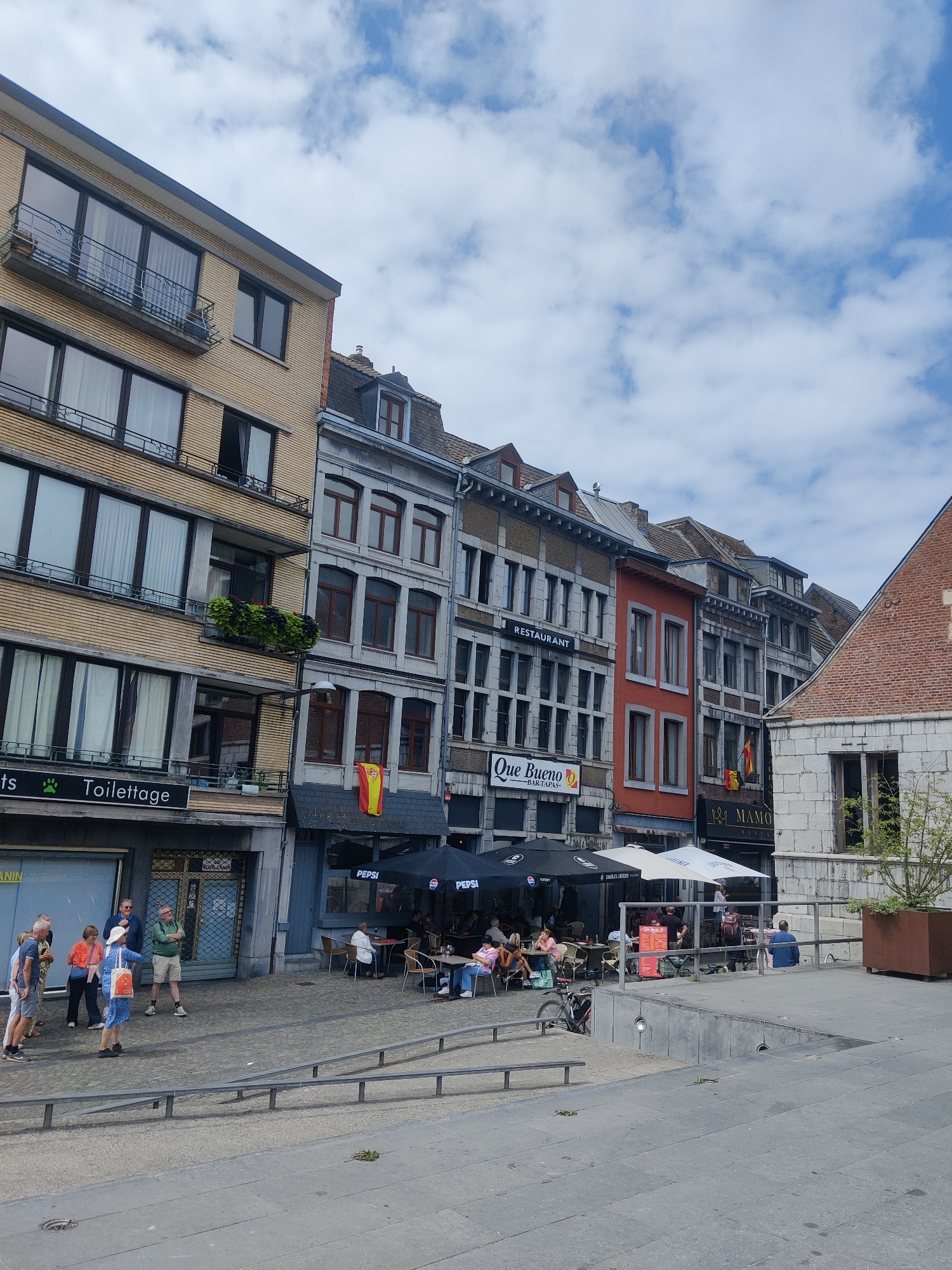 The image depicts a street scene in Liège, Belgium, with a focus on a row of buildings housing shops and restaurants. The architecture features a mix of modern and older styles, with the foreground showing a paved walkway and some people gathered.
In the center of the image, a building with a yellow brick facade stands out, featuring multiple stories with windows and balconies. Adjacent to it, other buildings display a more traditional European architectural style with stone or grey facades and multiple windows. Several signs are visible on the ground floor businesses, including "Toiletage" (pet grooming), "Que Bueno - Bar Tapas," and "Mamo - Restaurant." Umbrellas from outdoor seating areas of these establishments are also prominent.
Several individuals are visible in the scene. On the left side, a group of people are standing and conversing. In front of the restaurants, patrons are seated at tables, some engaged in conversation, others appearing to eat or drink. A bicycle is parked near one of the outdoor seating areas.
The environment includes a cloudy sky, suggesting overcast or partly cloudy weather. The street is paved, and metal railings and planters are part of the urban landscape. The overall impression is a moderately busy urban setting on what appears to be a daytime. The presence of businesses with outdoor seating indicates a public space conducive to socializing and dining.