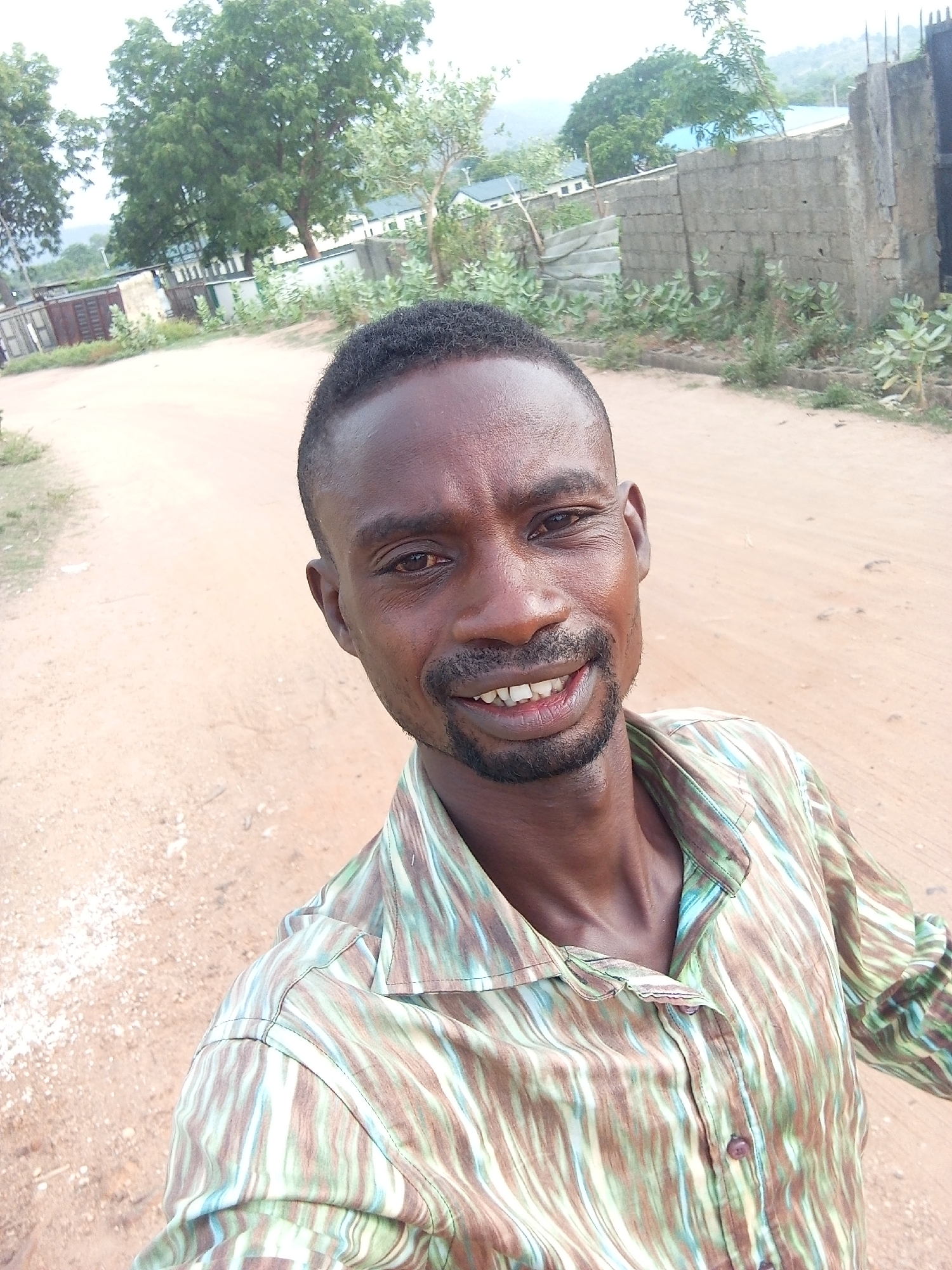 A man stands on a dirt road in Otokiti, Nigeria. He faces the camera and smiles. The dirt road has patches of exposed aggregate. A concrete block wall runs along one side of the road, with sparse vegetation growing near the base. In the background, trees, buildings, and a hill can be seen.