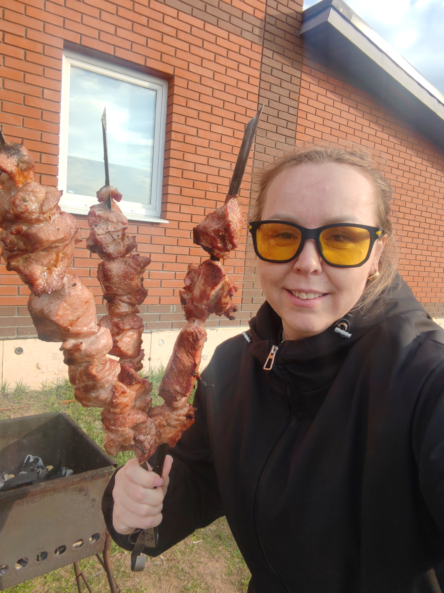 The image is a selfie of a person holding two skewers of cooked meat in front of a brick building with a window. The person is wearing yellow-tinted sunglasses and a black jacket. A charcoal grill is visible in the lower left of the image. The building is likely located in the city of Chaikovsky, Russia.