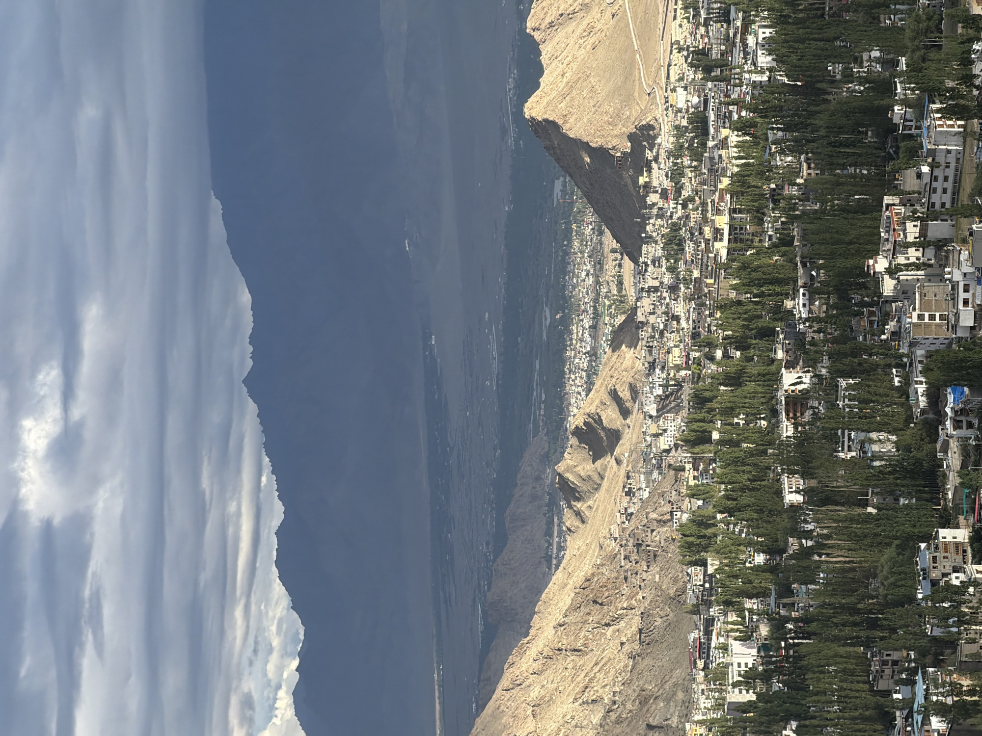 This is a high-angle, wide shot of Leh, India, bathed in the soft, diffused light of an overcast day. The sky is a dramatic expanse of layered grey clouds, with patches of brighter light suggesting breaks in the cloud cover. The background features the imposing, dark blue silhouettes of a vast mountain range, their jagged peaks softened by distance and atmospheric haze.
In the mid-ground, the arid, rocky slopes of the Himalayas dominate the landscape, their golden-brown hues catching the available light. Nestled within the valleys and spreading across the lower slopes is the city of Leh itself. Buildings, predominantly constructed with light-colored materials, are tightly packed together, creating a textured urban sprawl. The city is dotted with numerous tall, slender green trees, their dense foliage creating a striking contrast with the surrounding arid terrain. These trees are scattered throughout the urban area, lining streets and interspersed among buildings.
The foreground shows a closer view of the city's architecture, revealing various buildings of different sizes and designs. Some structures appear to be multi-story residential or commercial buildings, characterized by flat roofs and numerous windows. Evidence of infrastructure, such as roads and pathways, can be seen weaving through the city and up the hillsides. The overall impression is one of a densely populated settlement situated within a stark, mountainous environment. There are no visible people or active human behaviors depicted in the scene. The scene appears static and observational.