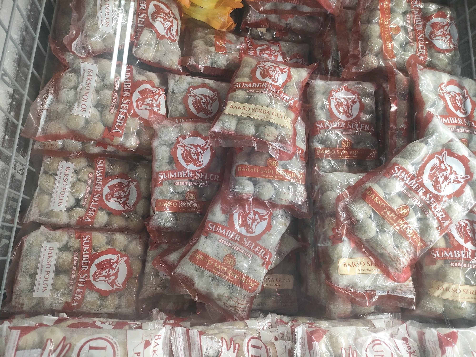 The image shows a freezer display case filled with multiple packages of frozen meatballs. The packaging is primarily clear plastic with red and white branding. Several labels are visible, including "SUMBER SELERA," "BAKSO POLOS," and "BAKSO SAPI PREMIUM." Some packages also feature a red circular logo with a bull's head. The meatballs themselves appear small and round, coated in a light-colored batter or ice. The freezer compartment is visible in the background, with a wire rack. The scene is typical of a supermarket or minimarket in Batam, Indonesia, where such frozen food products are commonly sold. No people are visible in the image.