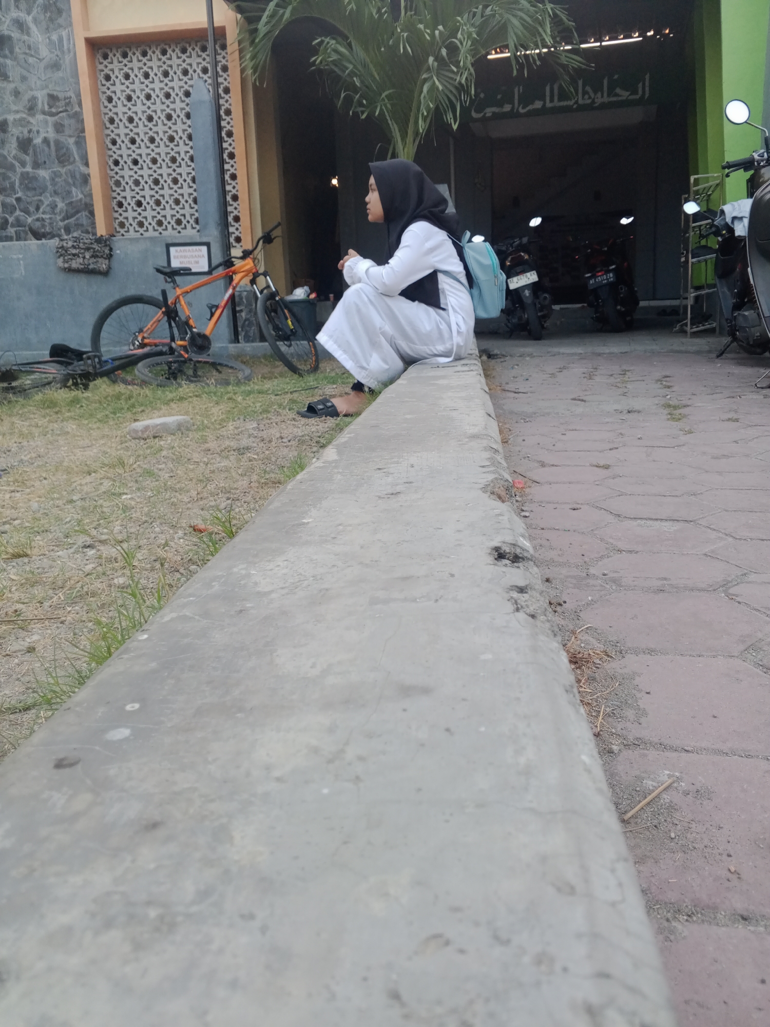 A young woman wearing a black hijab and white clothing is sitting on a concrete ledge, facing to the right. She has a light blue backpack next to her. In the background, there are two parked motorcycles and a bicycle leaning against a building. The building has grey stone walls and an orange-framed window. A sign with Arabic script is visible above a doorway. The ground is covered with gravel and grass.
