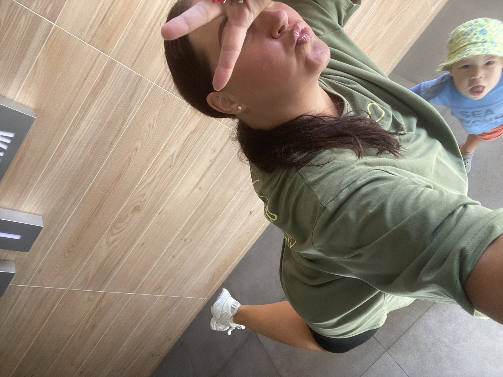 A woman in a green t-shirt and shorts is taking a selfie in a modern building with light wood-paneled walls. She is holding up two fingers to her face, covering one eye, and puckering her lips. Her right leg is extended behind her, showing a white sneaker and athletic shorts. To the lower right, a toddler in a blue t-shirt and a yellow patterned hat is visible. The floor is gray tile. The environment appears to be an indoor public space, possibly a mall or a building with a reception area, given the sleek wooden walls and the presence of signage that appears to be a part of the building's design. The overall impression is casual and domestic, with the woman capturing a moment with her child. There is no indication of the specific location within Sochi, Russia, beyond it being an indoor public setting.