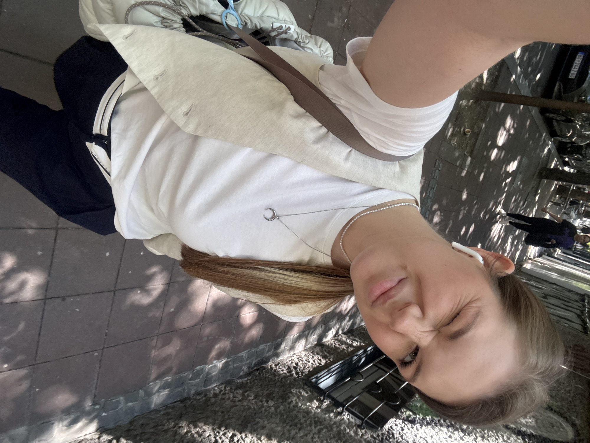 A woman is taking a selfie on a city street. She wears a white t-shirt, a tan blazer, dark pants, and a crossbody bag. She is wearing a necklace and an AirPod in her right ear. She is squinting one eye. The location is Belgrade, Serbia, as indicated by the license plate on a car in the background. There are pedestrians in the distance.