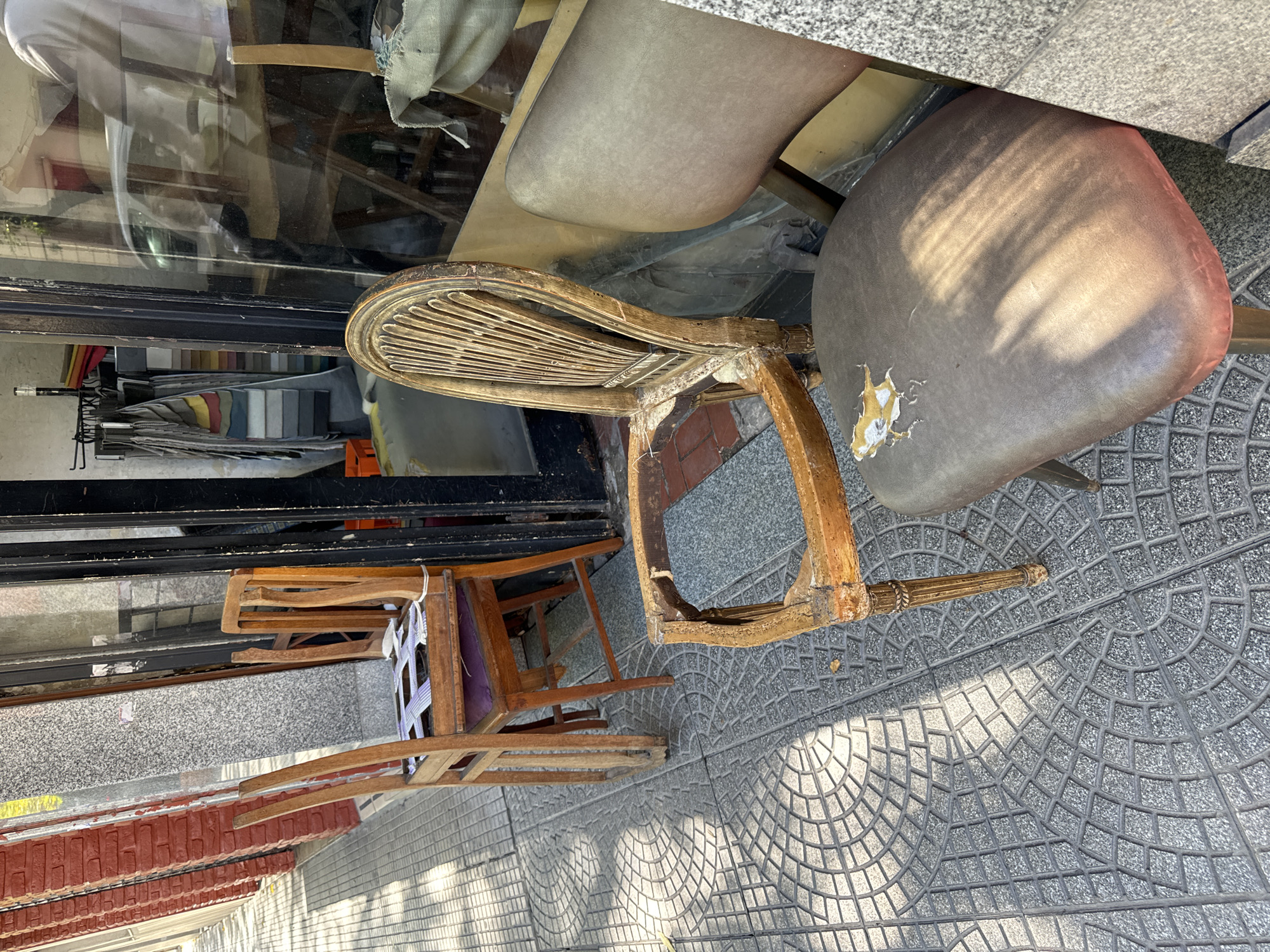 This image, captured in Buenos Aires, Argentina, presents an outdoor scene featuring an array of old and worn chairs situated on a patterned sidewalk outside what appears to be a furniture or upholstery shop.
In the foreground, a prominent wooden chair with a distinctive slatted, curved back is heavily damaged, with a significant portion of its seat and frame broken away. Adjacent to it is another chair, its dark, possibly faux leather, upholstered seat showing considerable wear and tear, with several tears exposing the lighter material beneath. Part of a decorative leg from this chair is visible on the sidewalk. Further inside the scene, closer to the shop window, additional chairs can be seen, including one with a purple cushion and another with a woven seat, suggesting a collection of items either for repair, display, or disposal.
The shop window, partially reflective, reveals an interior displaying numerous fabric swatches or samples in various colors and textures, along with some umbrellas, indicating a business specializing in textiles or furniture restoration.
The scene is bathed in natural daylight, with sunlight casting distinct shadows of the chairs and the shop's structure onto the sidewalk. The grey sidewalk tiles feature an intricate pattern of interlocking square and radial designs, typical of urban environments in Buenos Aires. The time of day appears to be daytime, likely mid-morning or afternoon, judging by the length and direction of the shadows. No individuals are present in the image, and no decipherable text is visible. The overall mood suggests a collection of forgotten or soon-to-be-restored furniture.
