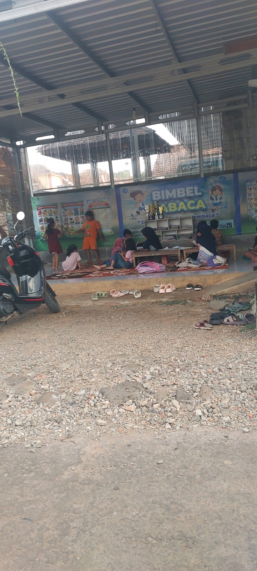 The image depicts an outdoor scene in Keling, Indonesia, likely an informal learning or community space. In the background, a mural with colorful illustrations and text, including "BIMBEL MEMBACA," suggests a reading or tutoring center. Several children are present. Two girls in the foreground are playing on a mat, one in a red dress and the other in an orange t-shirt and shorts. Another child in a pink shirt is sitting on the mat. Further back, two individuals in headscarves are seated at a low wooden table, appearing to be interacting with books or other materials. A shelf unit filled with items is visible behind them. Several pairs of sandals and shoes are scattered on the ground in front of the shaded area. A motorcycle is parked to the left of the frame. The ground in the foreground is rough and covered with gravel and small stones. The overall environment appears to be a covered outdoor area with a corrugated metal roof supported by visible beams.
