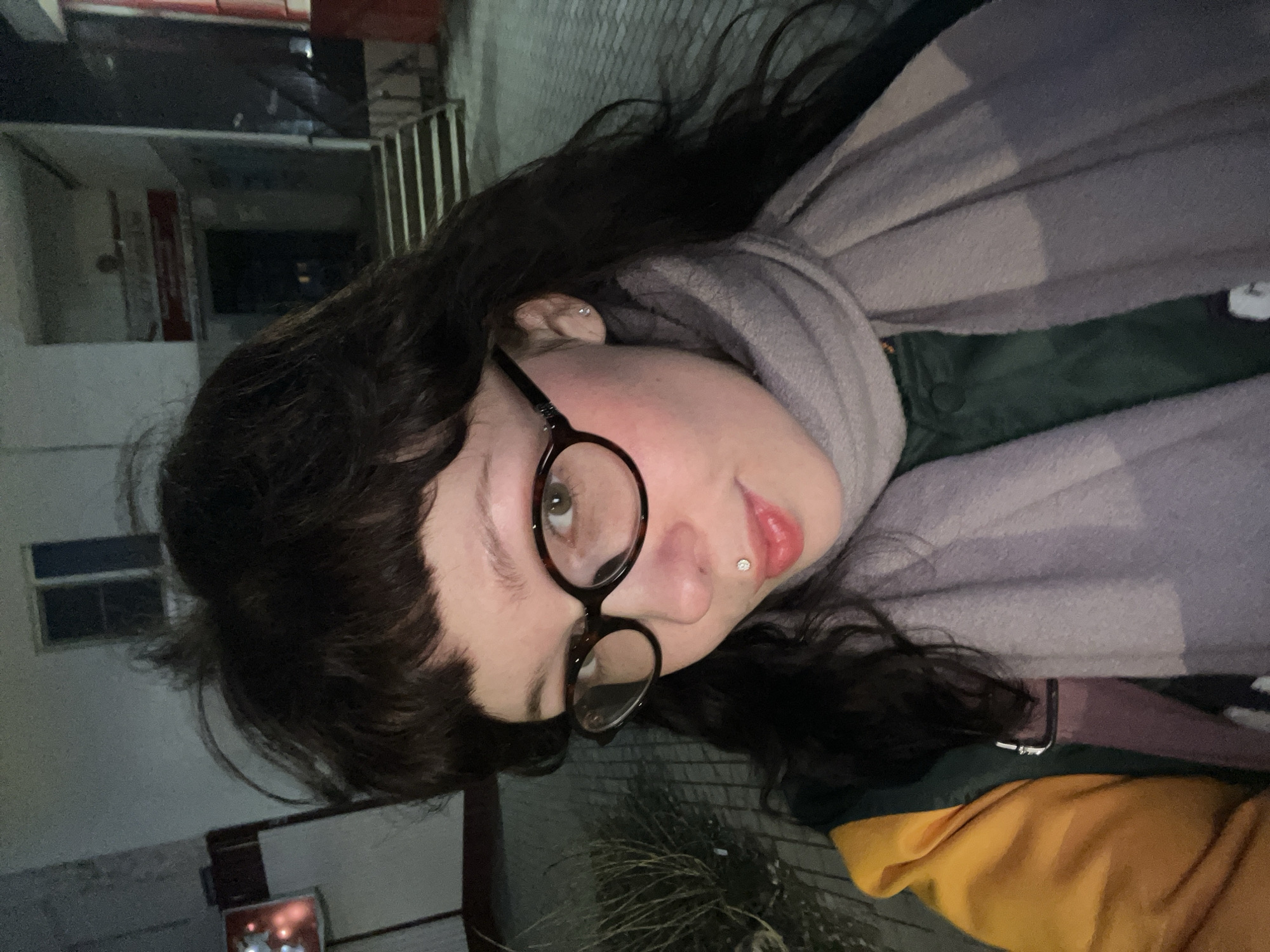 The image displays a selfie of a person, likely female, captured outdoors at night or dusk in Chaikovsky, Russia. The individual has dark, wavy hair with bangs and wears distinctive round, tortoise-shell glasses that frame light-colored eyes, appearing green or hazel. A small, sparkling stud is visible in a labret piercing positioned centrally below the lower lip, which is adorned with light pink lipstick. A slight rosy flush is present on the cheek, and a small stud earring can be seen in the visible ear.
The person is dressed warmly, indicative of cool or cold weather. They are wearing a thick, light grey or beige scarf wrapped around their neck and shoulders. Beneath the scarf, a dark green top or jacket is visible, along with a bright mustard yellow garment layered underneath.
The background is dimly lit by artificial light sources, consistent with a nocturnal setting. It features indistinct urban or semi-urban elements, including parts of buildings with windows and a metallic railing, suggesting a street or pedestrian area. Sparse, dry vegetation is visible in the lower foreground. The overall atmosphere is casual, and the person's expression appears neutral or subtly contemplative. No discernible text is visible in the image.
