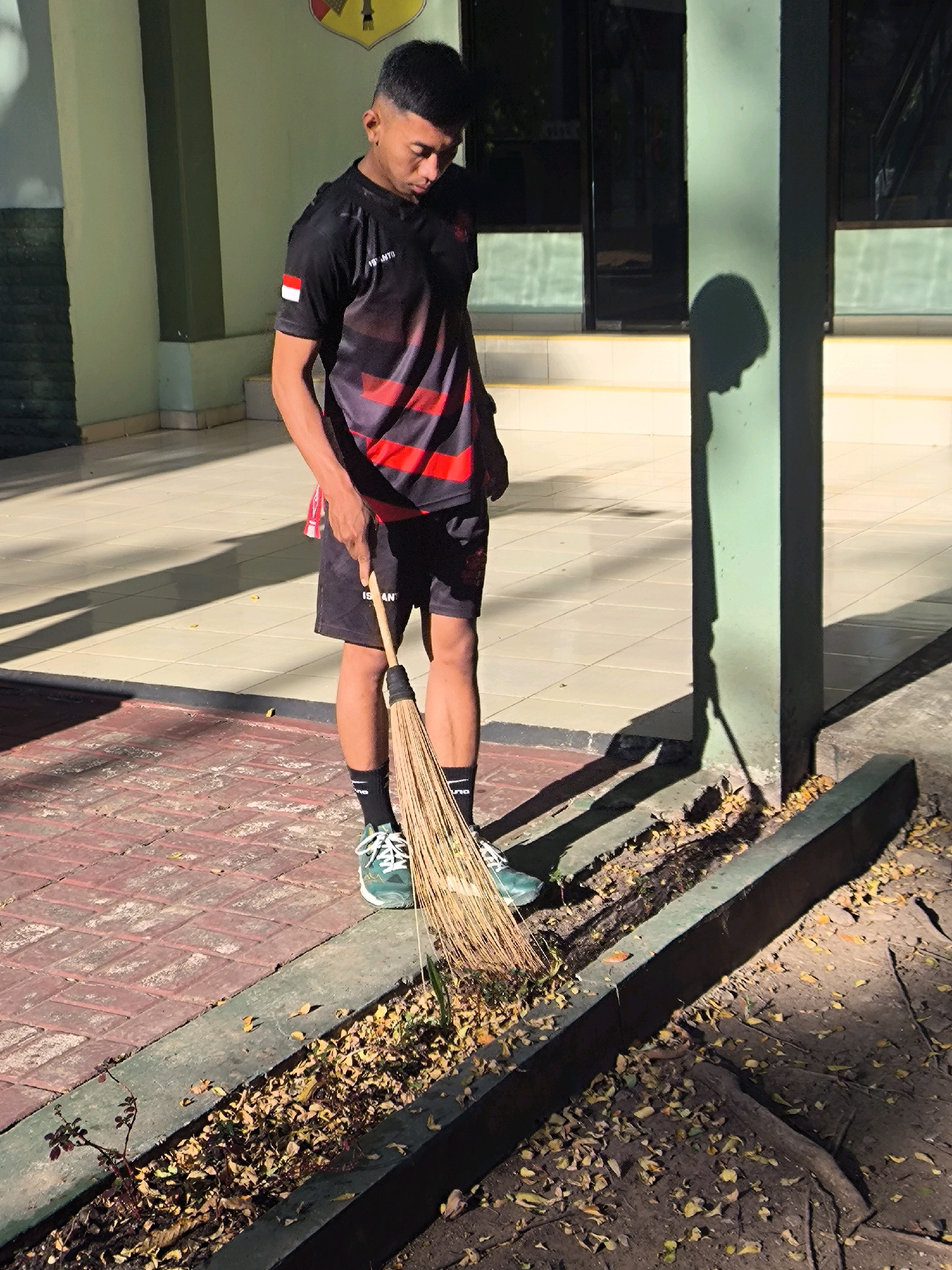 A young male, appearing to be of Southeast Asian descent, is shown standing on a paved walkway and is in the process of sweeping. He is wearing a black t-shirt with red stripes and black shorts, black socks, and green athletic shoes. He holds a broom made of straw with a wooden handle in his right hand and is sweeping leaves from a raised garden bed. The background includes a light green wall, a building with windows, and tiled flooring. The scene appears to be outdoors, possibly in a school or public area, given the architecture and the presence of the broom, suggesting a cleaning task. The lighting indicates it is daytime with strong shadows cast by the man and a nearby pillar. The image does not provide specific information about a location within a city in Indonesia, beyond its general appearance.